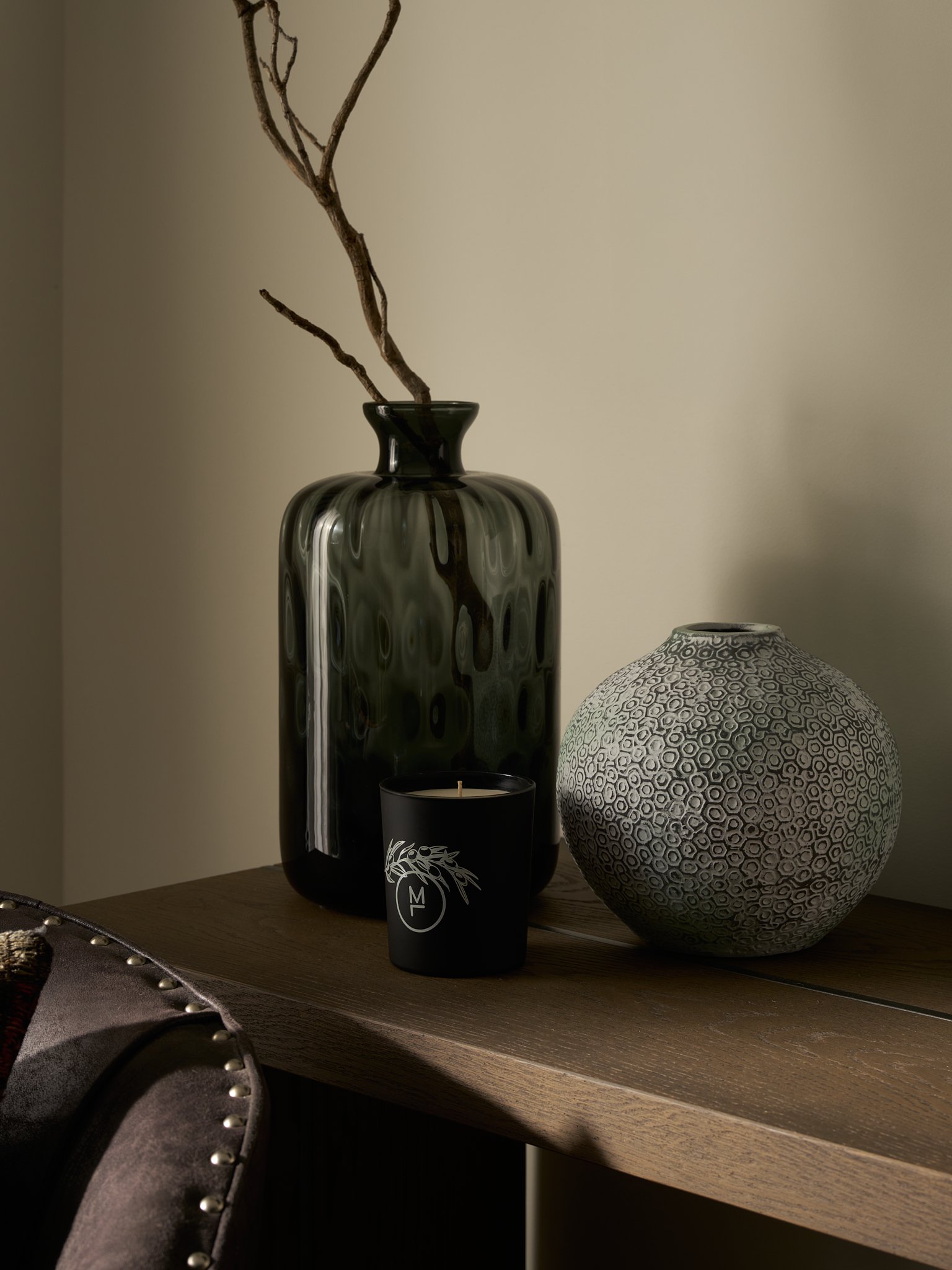A black and white vase with a branch inside on a wooden surface, with a decorative black candle holder featuring a white leaf pattern and a black candle with a white logo. To the right is a patterned, rounded ceramic vase on a wooden surface.