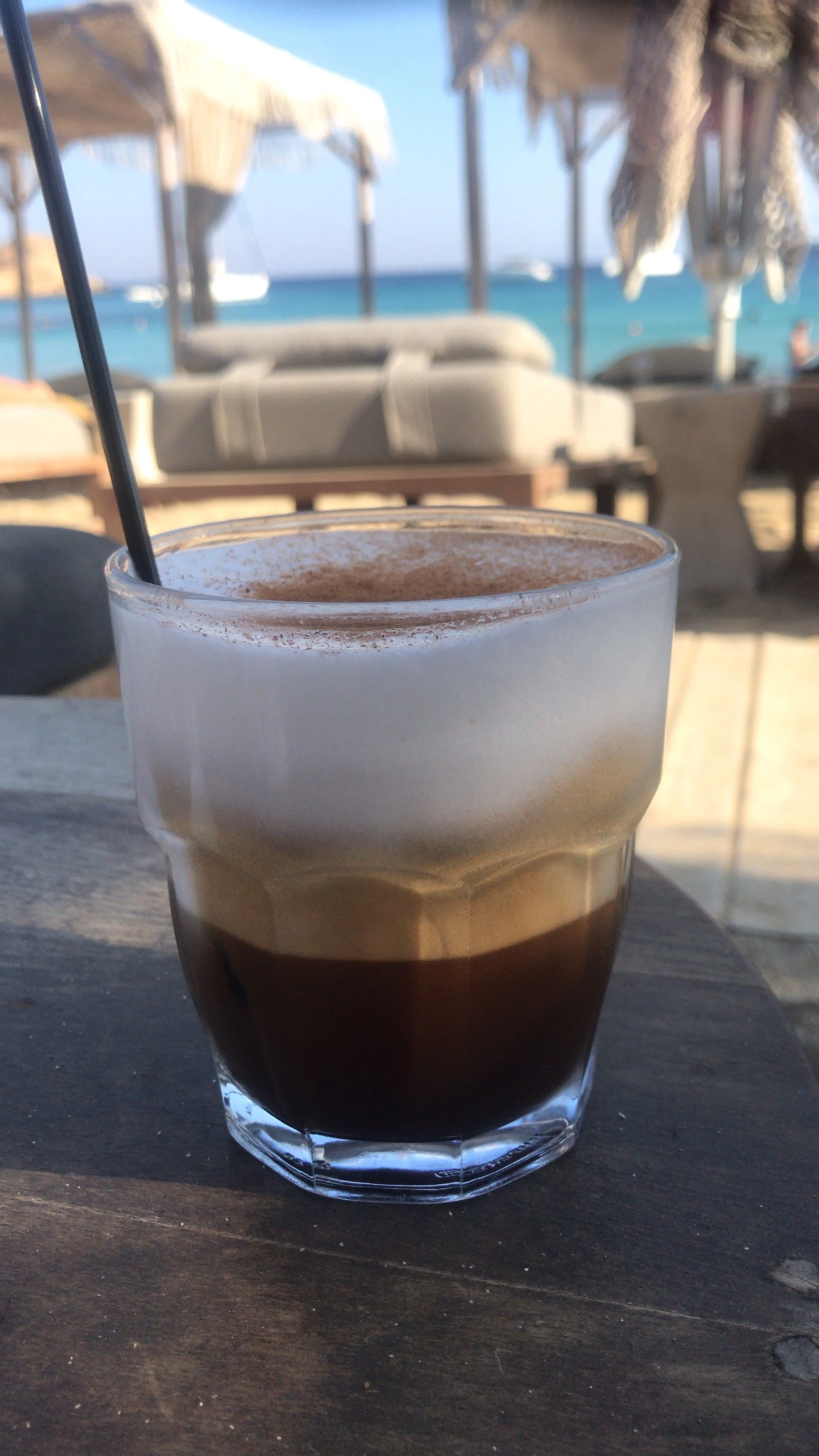 A glass of iced coffee with a black straw on a wooden table at the beach, with umbrellas, lounge chairs, and the ocean in the background.
