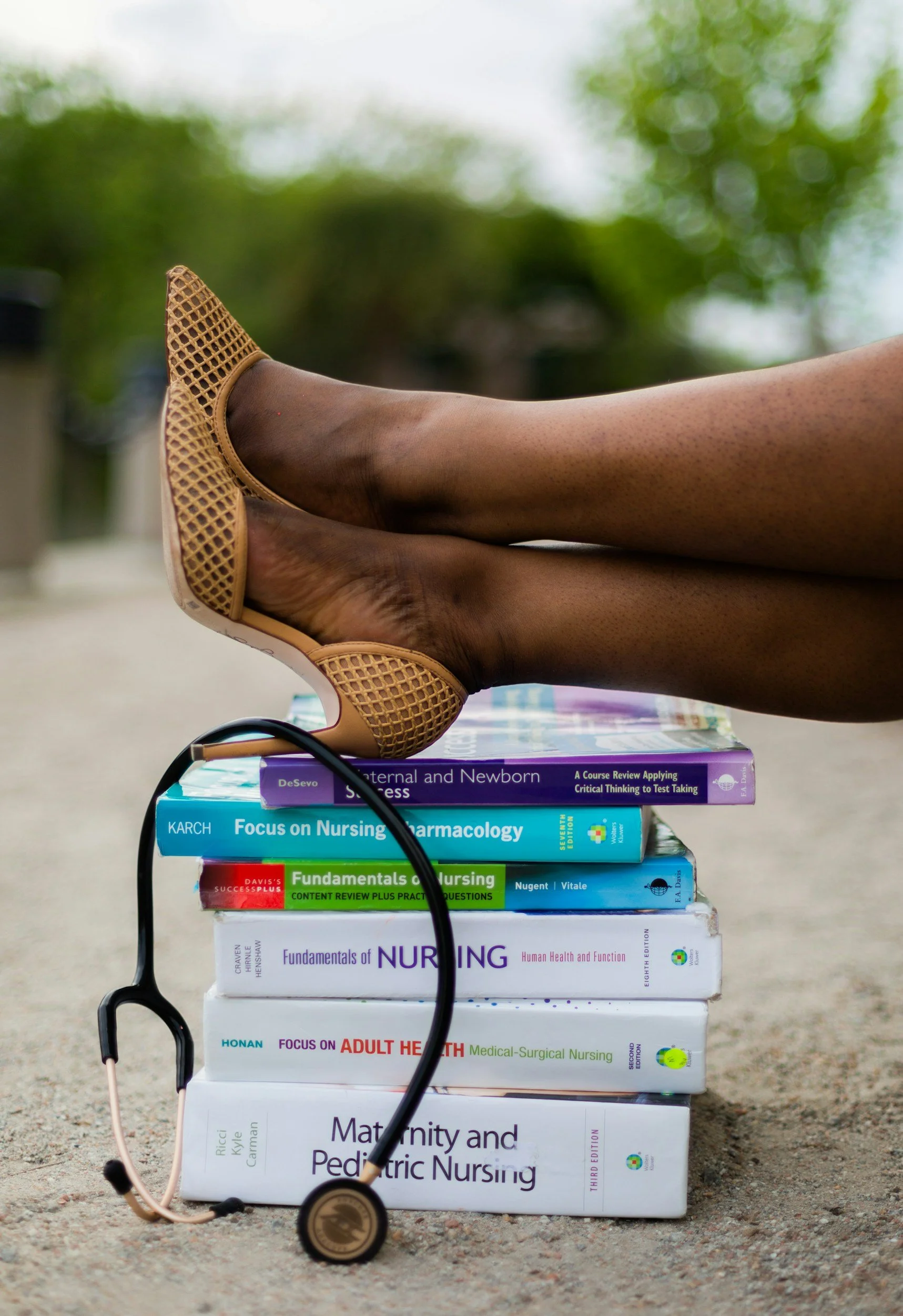 Stack of nursing textbooks with a stethoscope and a pair of shoes on top, outdoors with green trees in the background.