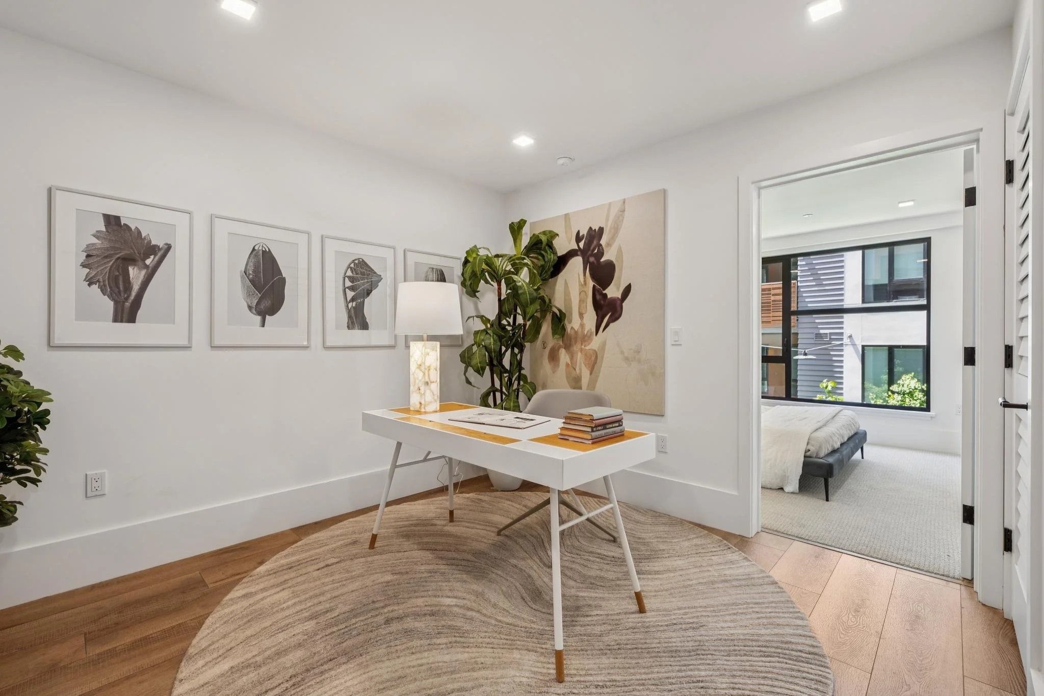 A modern interior room with white walls, a small white desk with scattered papers and books, a tall lamp, framed black-and-white flower photographs on the wall, a large abstract artwork, a potted plant, a wooden floor rug, and an open door leading to a bedroom with a bed and a window.