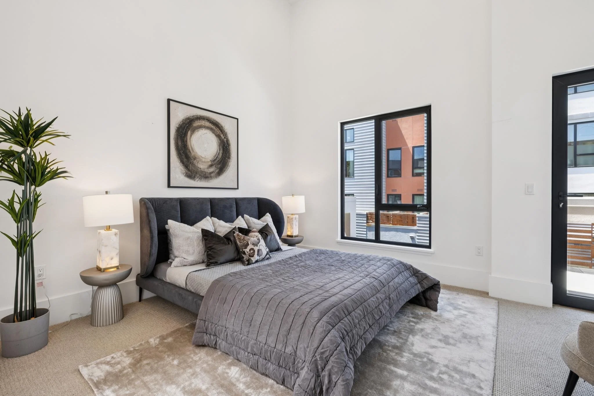 Modern bedroom with a gray upholstered bed, multiple pillows, abstract circular art on the white wall, two side tables with lamps, a tall green plant in a gray pot, and large windows showing building exteriors.