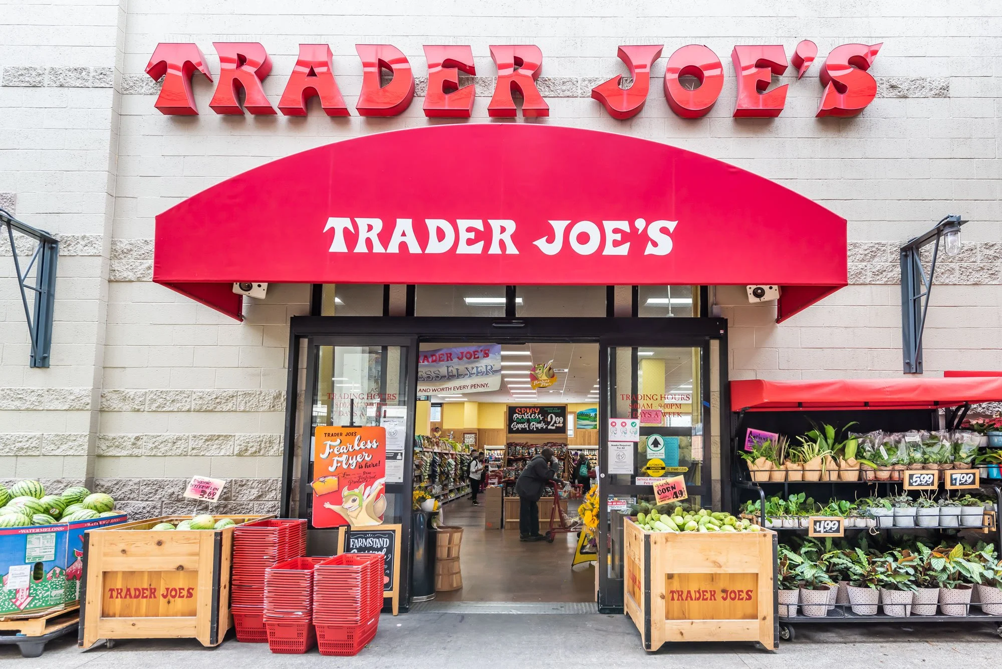 Exterior view of a Trader Joe's grocery store entrance, with a red awning displaying the store's name and various fresh produce like watermelons and plants outside.