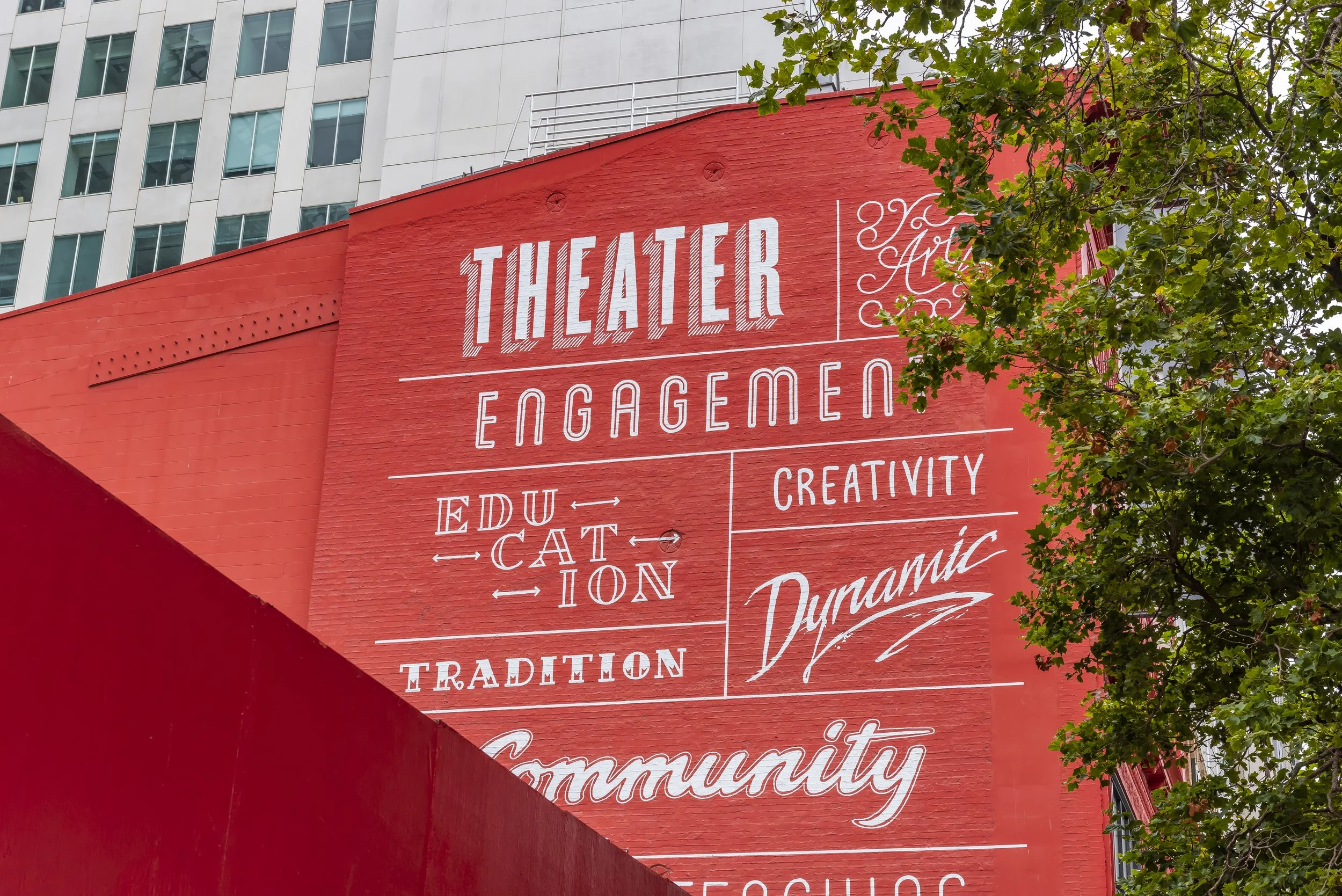 Red brick wall with white painted words highlighting themes like theater, education, creativity, tradition, and community, with a modern building and green tree leaves in the background.