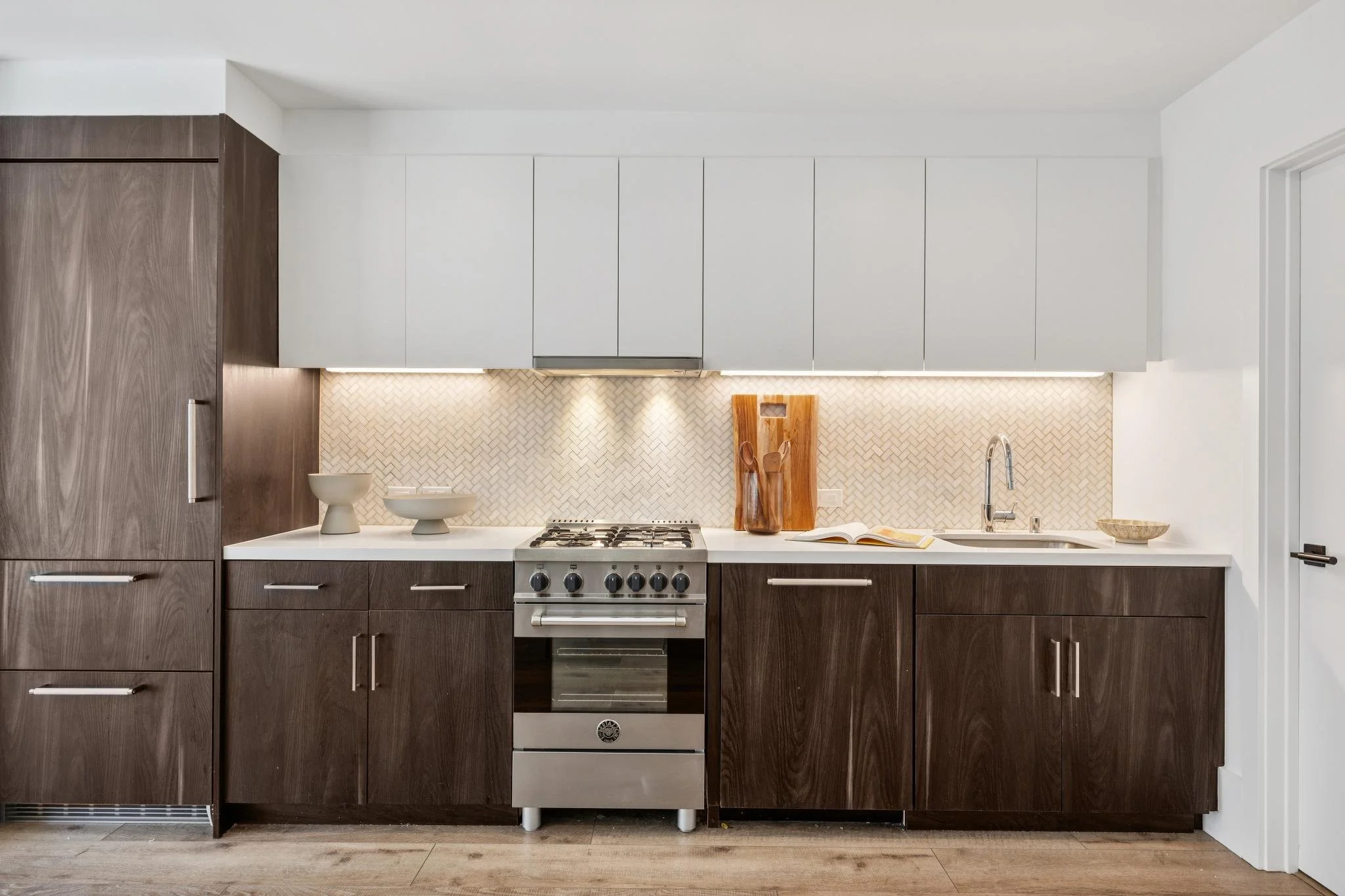 Modern kitchen with dark wood lower cabinets, white upper cabinets, stainless steel stove, beige herringbone backsplash, and wooden cutting board with utensils on the countertop.