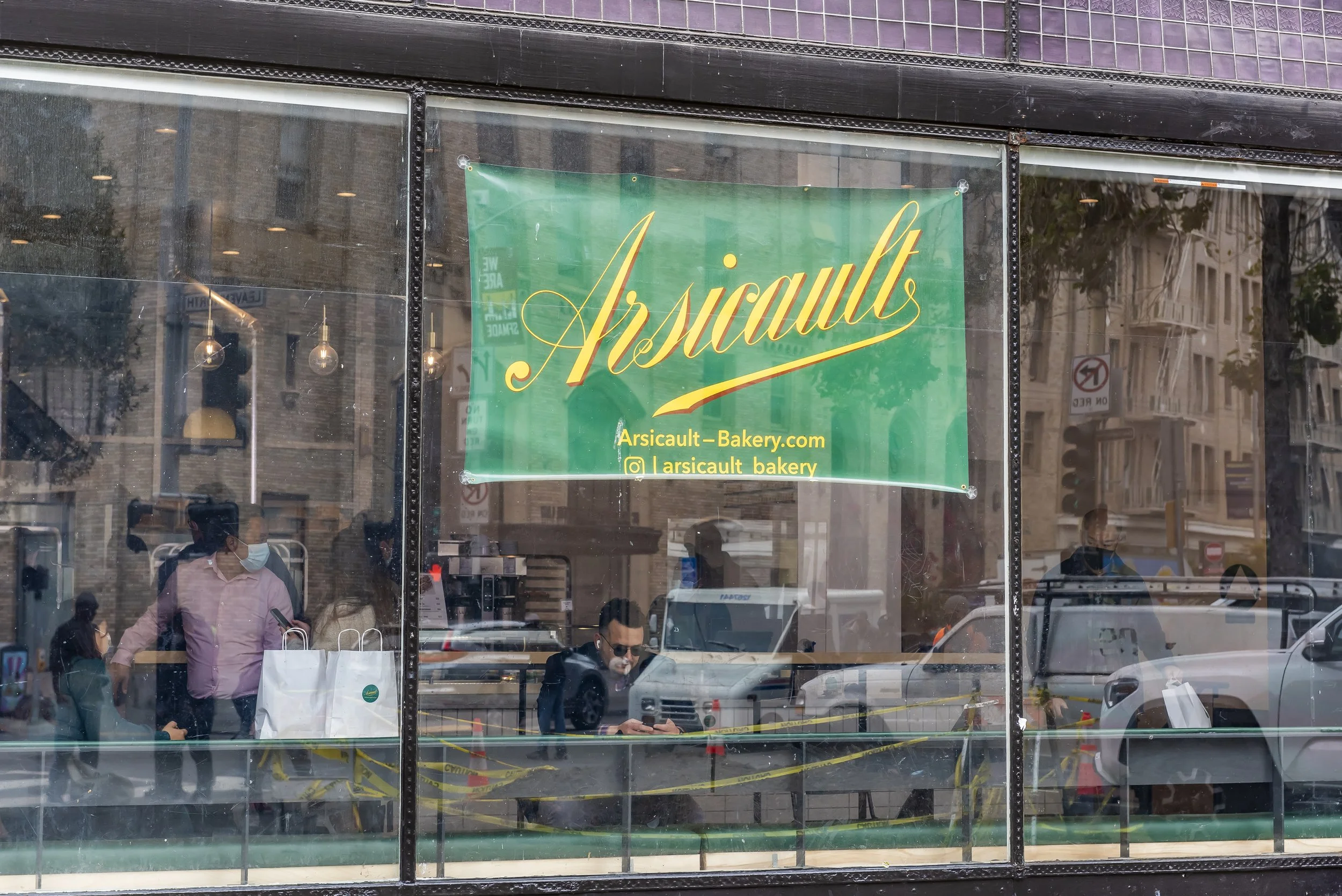 Exterior view of a bakery with a large green sign displaying the name 'Arsiqault' in yellow cursive font. The sign also includes the website 'Arsiqault-Bakery.com' and social media handle '@arsiqault_bakery'. Through the glass window, people can be s