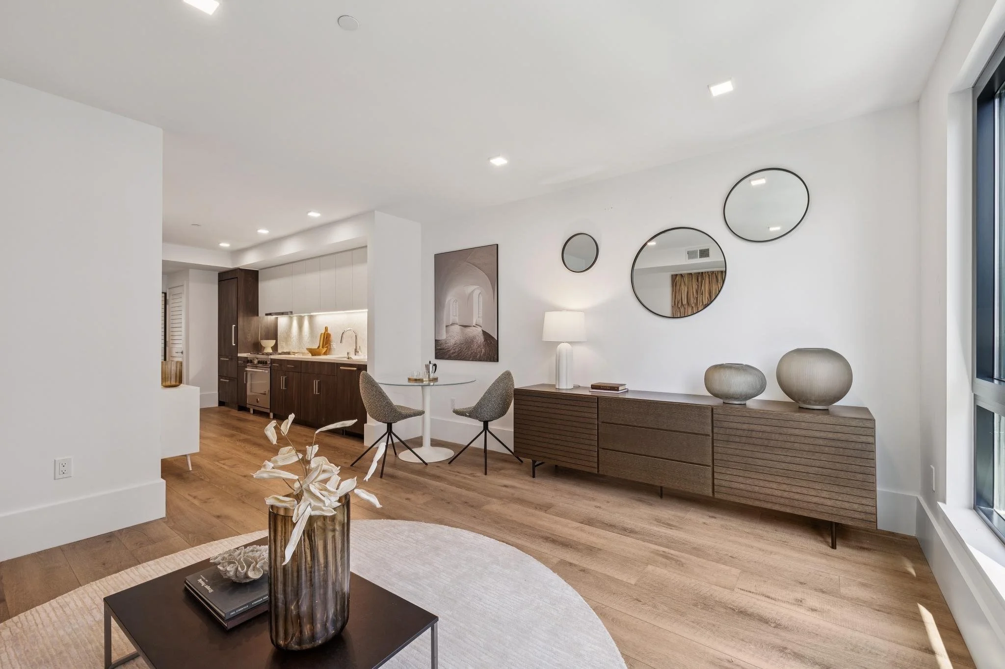 Modern living room with white walls, wooden flooring, and minimalistic decor. There is a black coffee table with a large gold and brown vase and decorative flowers. A beige rug is under the table. In the background, there are two gray swivel chairs, 