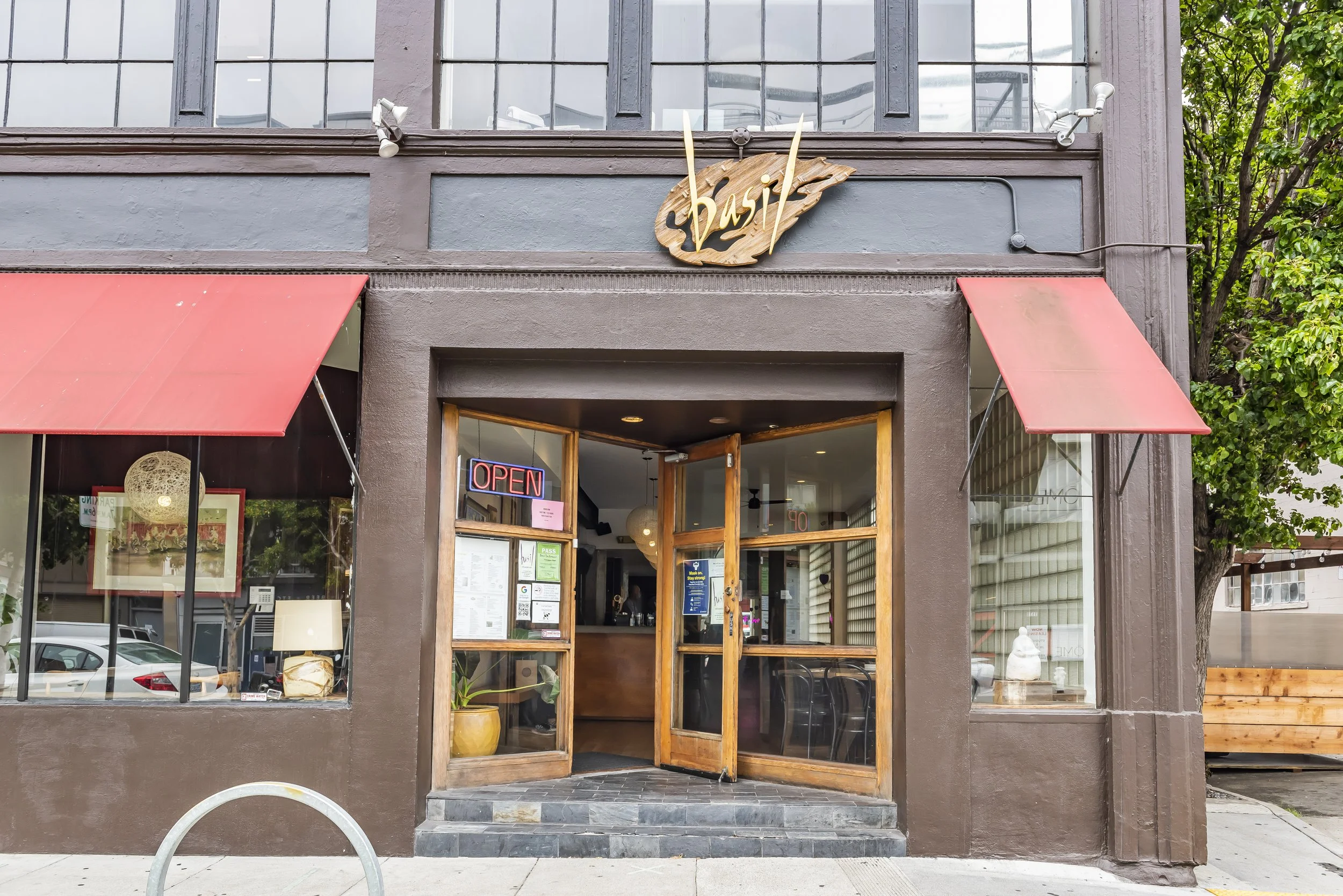 Front entrance of a restaurant with a sign that says 'basil'. The sign is styled as a leaf with cursive writing and two blades or leaves extending from the top. The entrance has wooden and glass doors with an 'Open' sign and menu visible inside. Two 