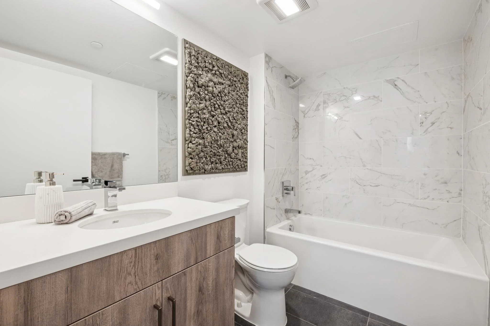 Modern bathroom with white marble walls, a bathtub, a toilet, and a vanity with a white countertop, mirror, and decorative items including a towel, soap dispenser, and a artwork on the wall.