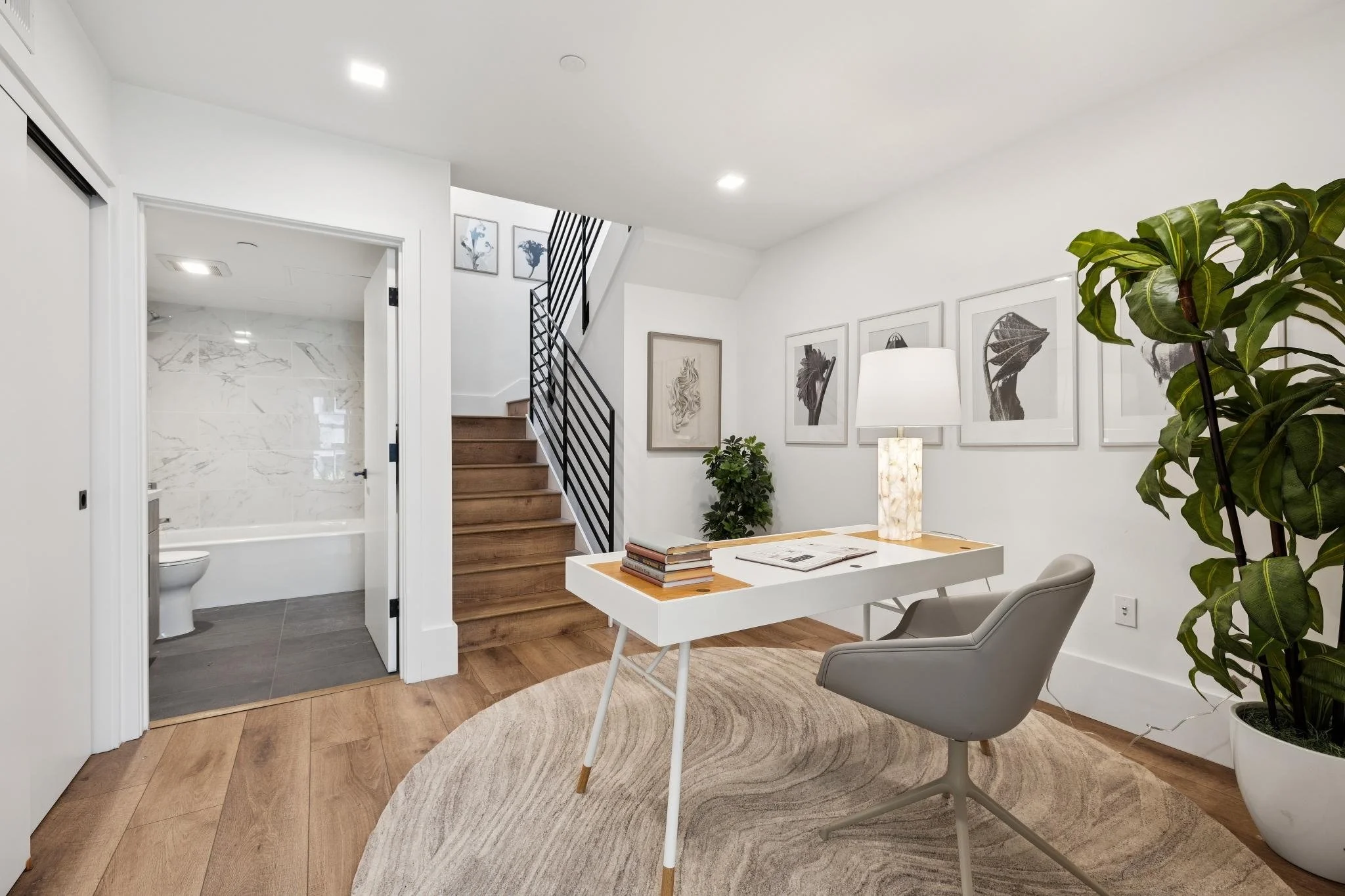 Modern interior of a living space with wooden flooring, a white desk with books and a lamp, a gray chair, framed artwork on white walls, large green plants, and a staircase leading upstairs. There is also an open doorway revealing a bathroom with mar