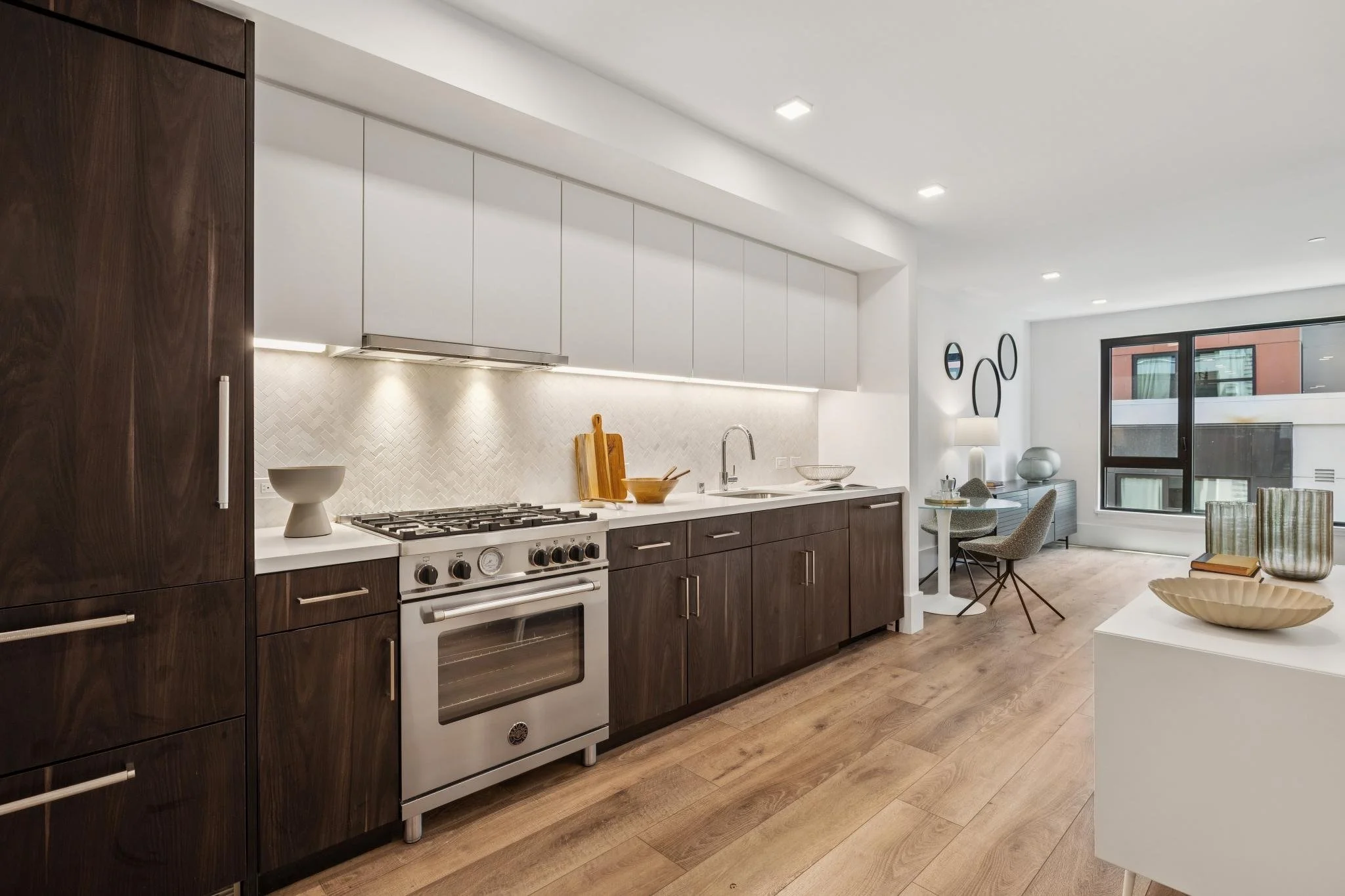 Modern kitchen with wood and white cabinets, stainless steel oven, and a view of the living area with a table, chairs, and decor.