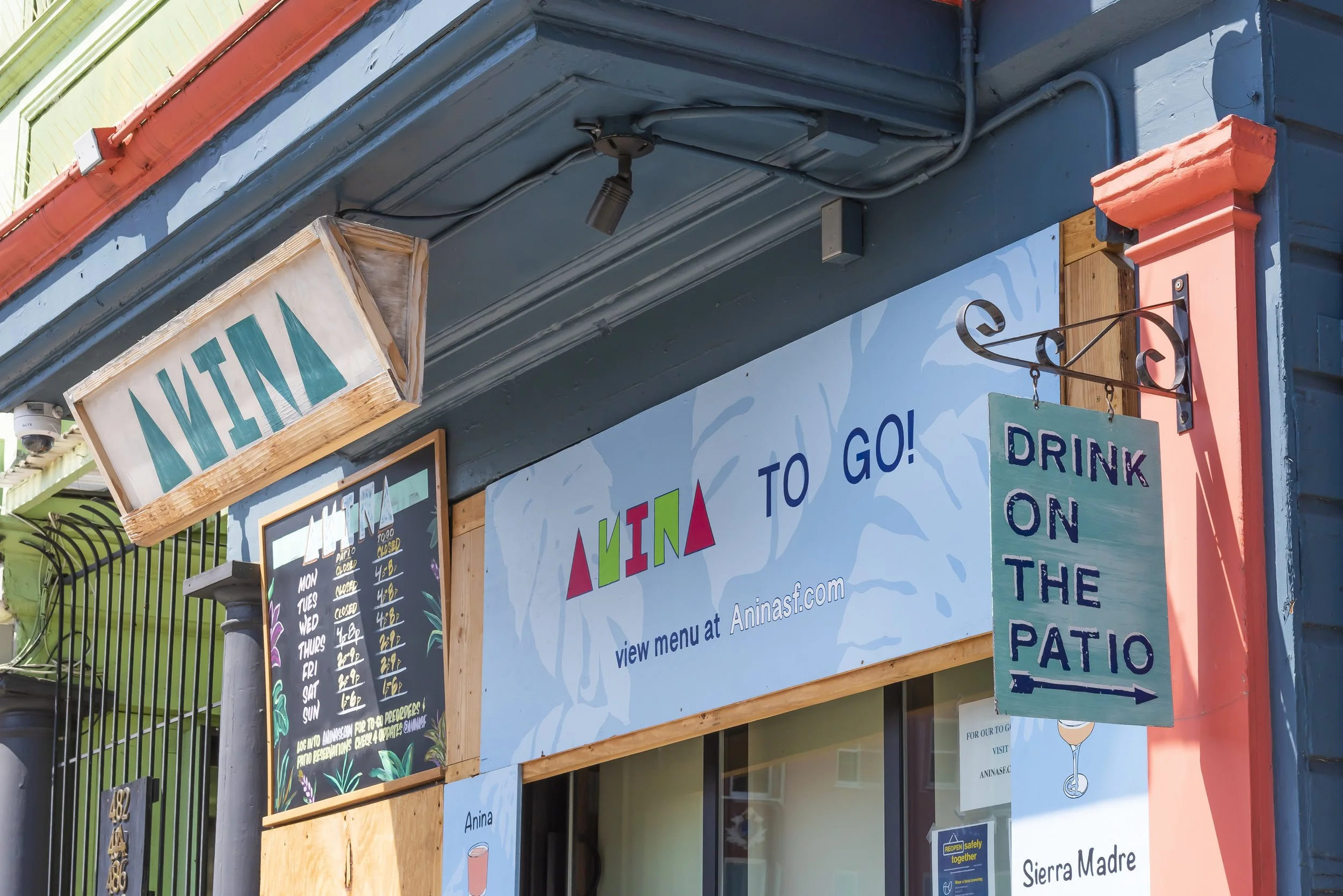 Colorful storefront with multiple signs including a wooden menu board, a hanging sign that reads 'Menu,' and a large blue and white sign promoting patio drinks at Anina. The image shows part of the building's exterior with blue, pink, and green accen