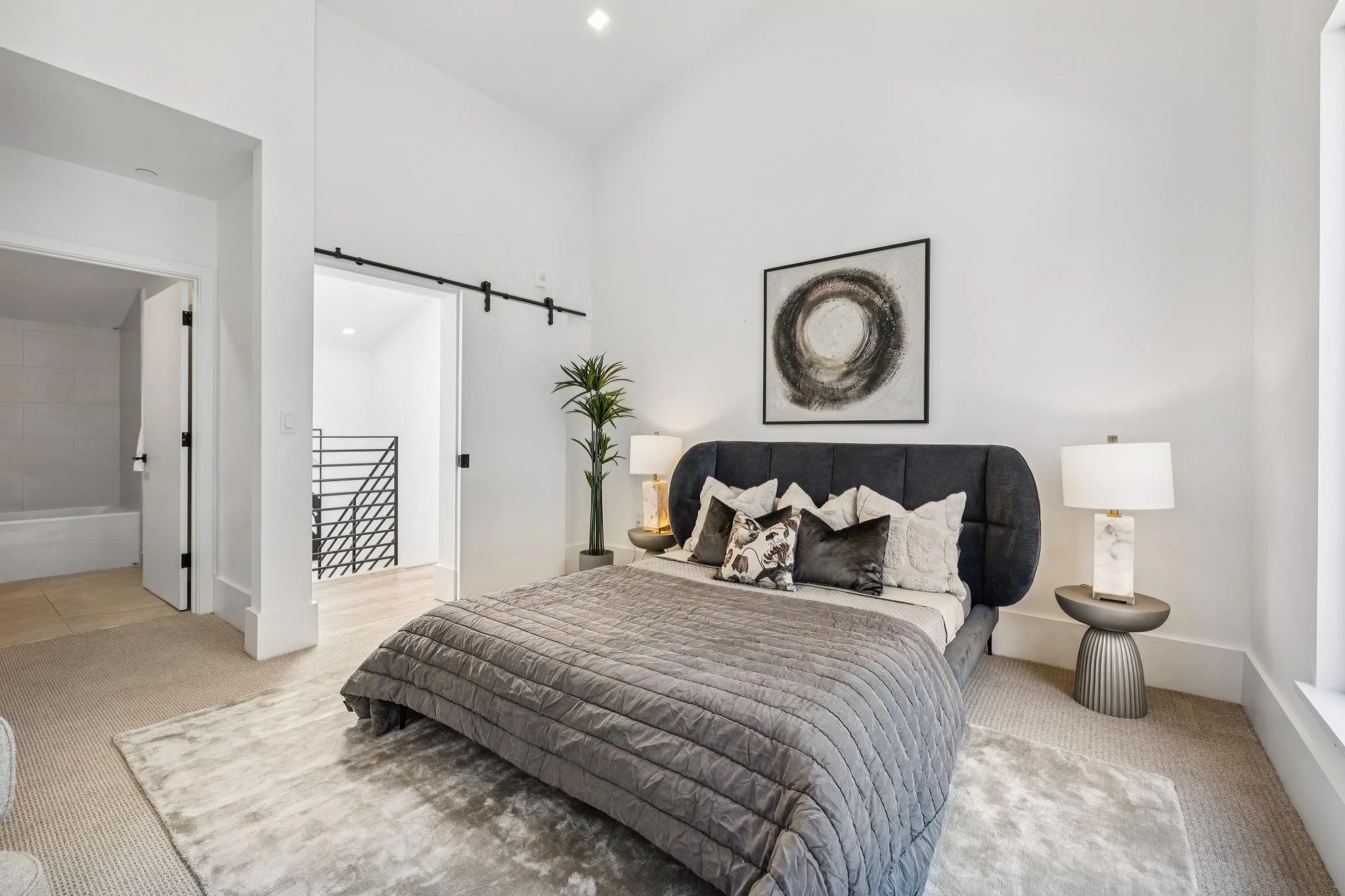 Modern bedroom with a gray upholstered bed, decorative pillows, white nightstands with lamps, a framed abstract circle artwork, a tall plant, and a sliding barn door leading to another room. The walls are white, and there is a beige area rug on light