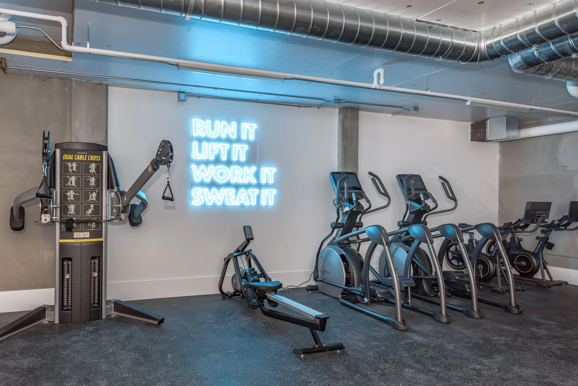 Fitness gym with rowing machines, stationary bikes, and an orange and black dual cable crossover machine. A neon sign on the wall reads, 'Run It Lift It Work It Sweat It.' The gym has an industrial style with exposed ductwork on the ceiling and a con