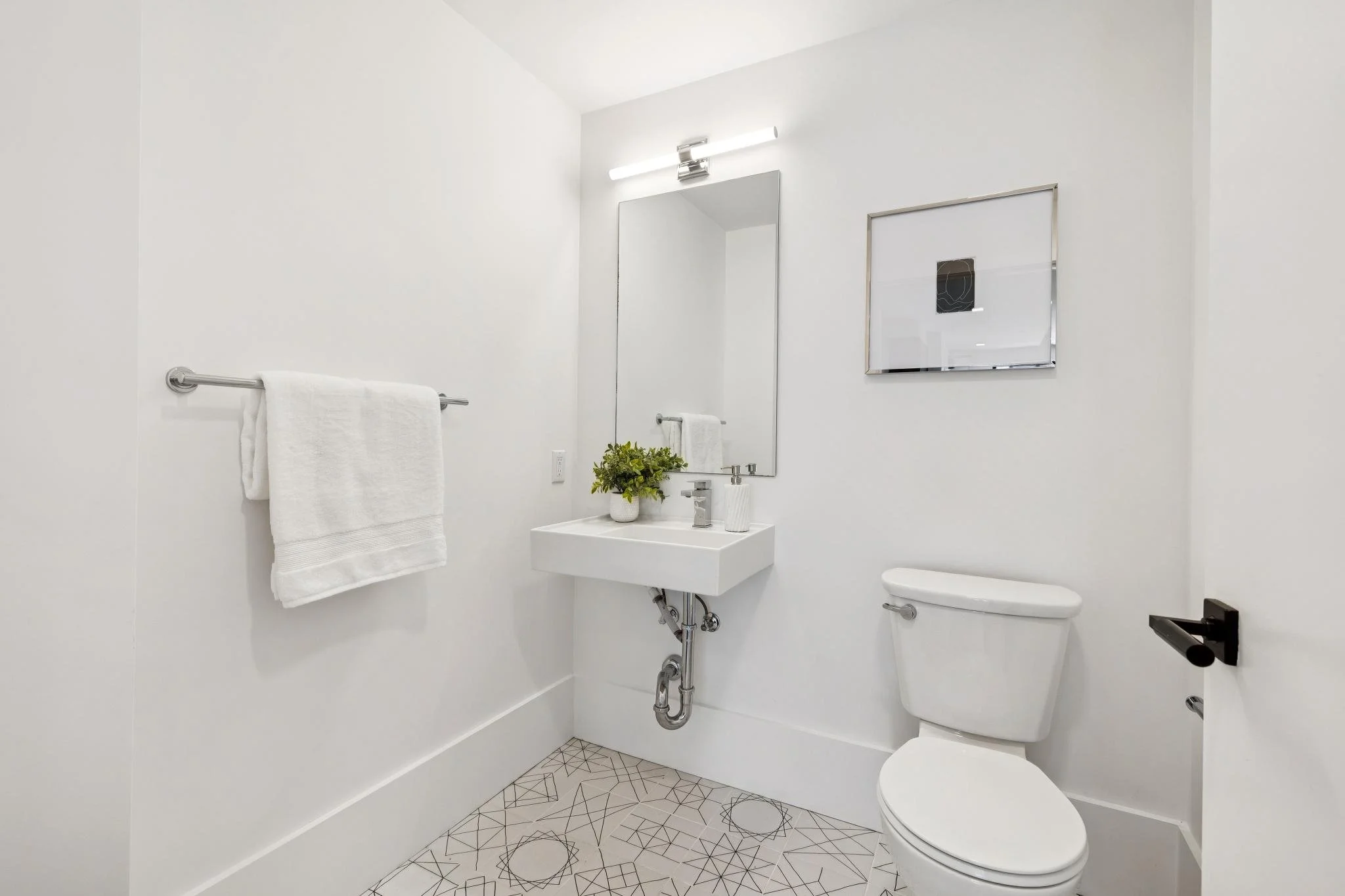 Minimalist bathroom with white walls, a small wall-mounted sink, a rectangular mirror, a towel rack with a white towel, a potted green plant, a toilet, and artwork on the wall. The floor has geometric patterned tiles.