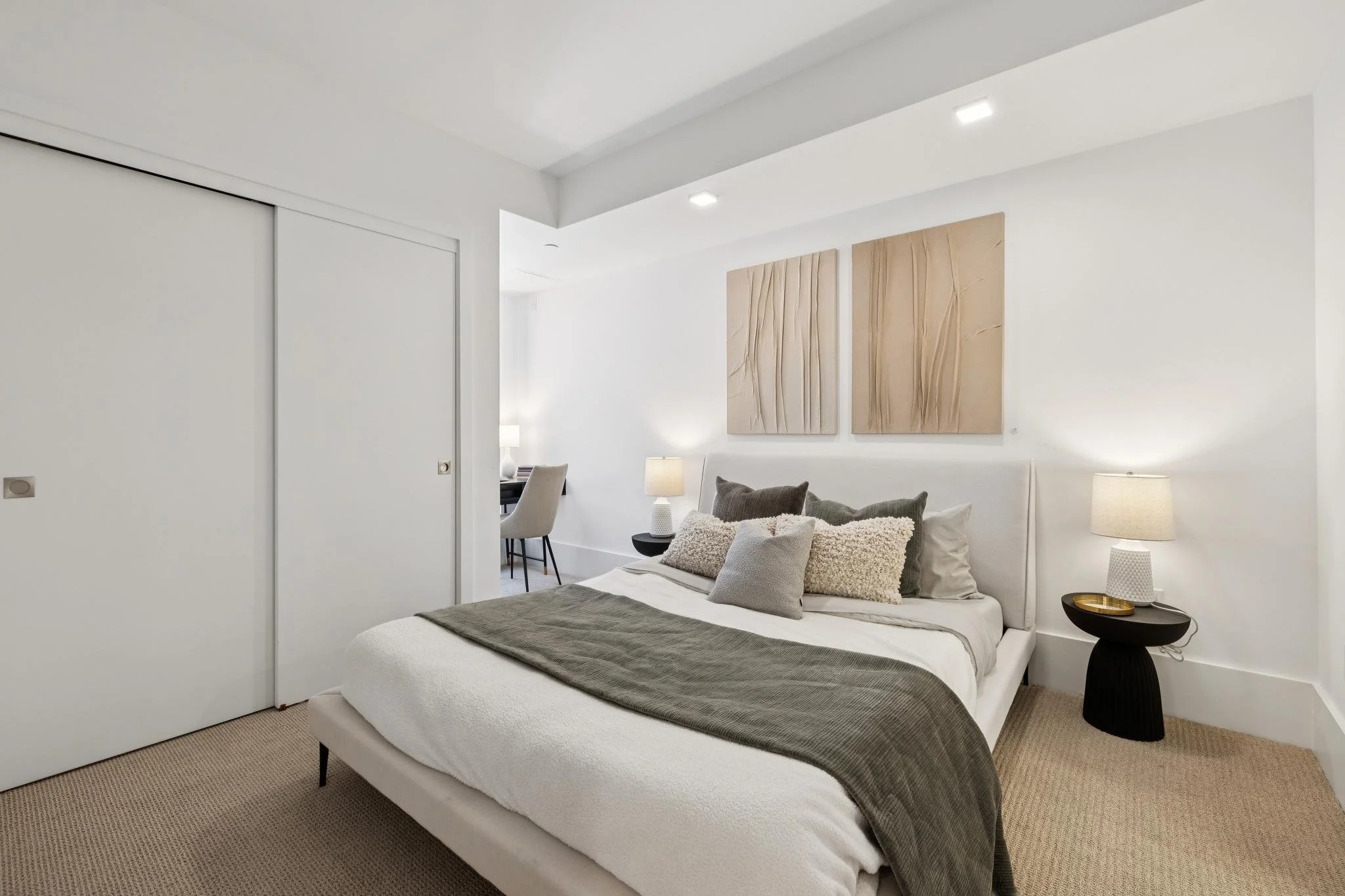 Modern bedroom with white walls, beige carpet, a bed with gray, white, and beige pillows, and a gray blanket. There are two large abstract art pieces above the bed, a black side table with a white textured lamp, and a small gold tray. A doorway on th