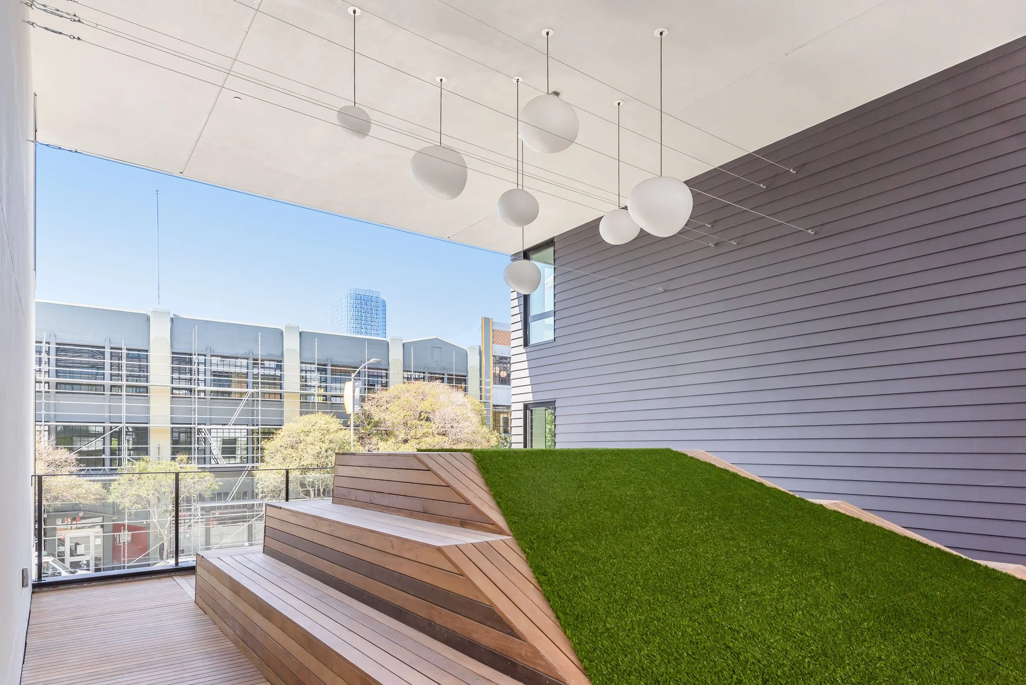 Indoor balcony with modern wooden benches and artificial grass, overlooking neighboring buildings and blue sky.