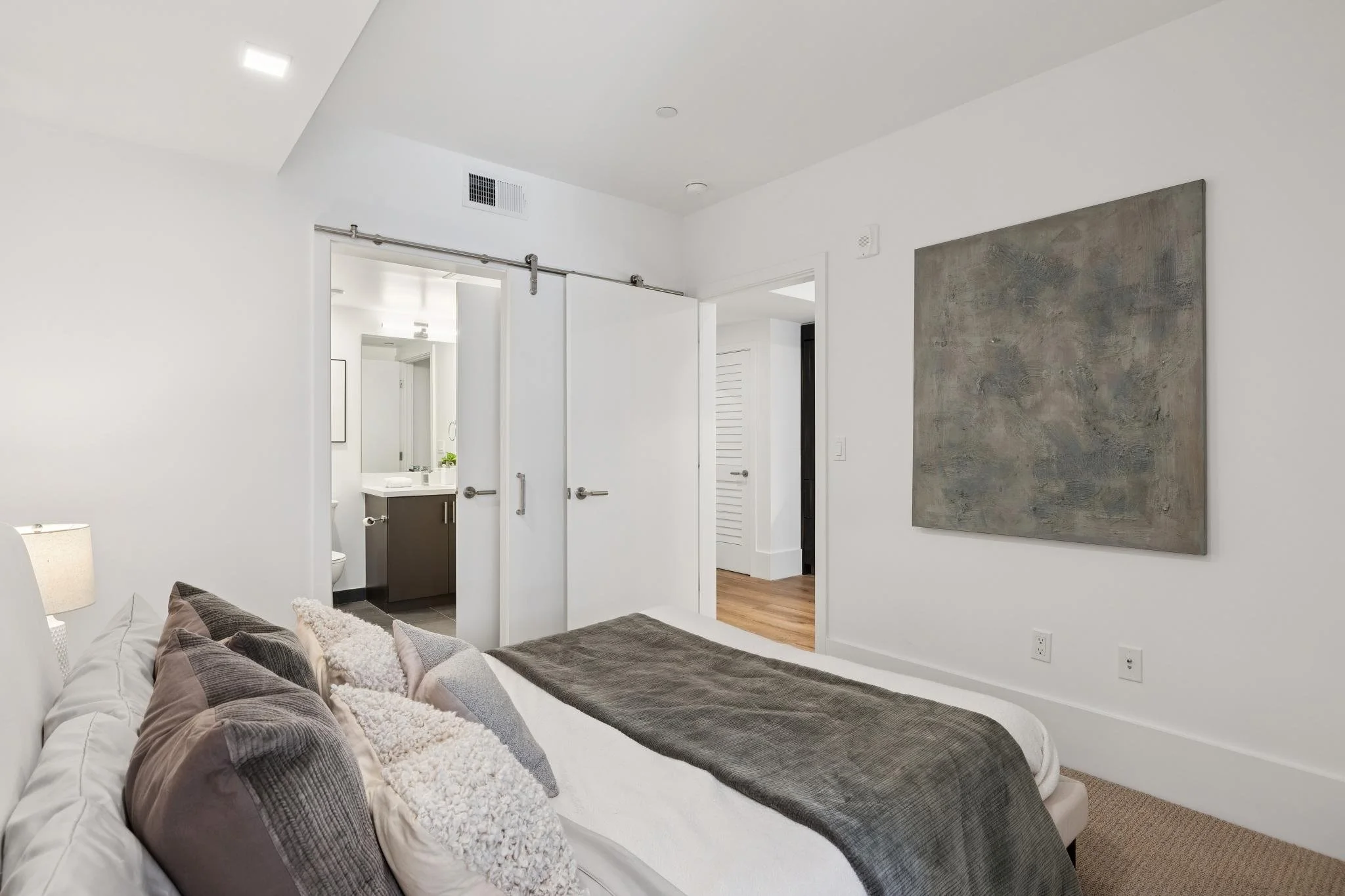 A modern bedroom with a bed that has multiple pillows and blankets. There is a painting on the wall and an open door leading to a bathroom with a sink, mirror, and toilet visible.
