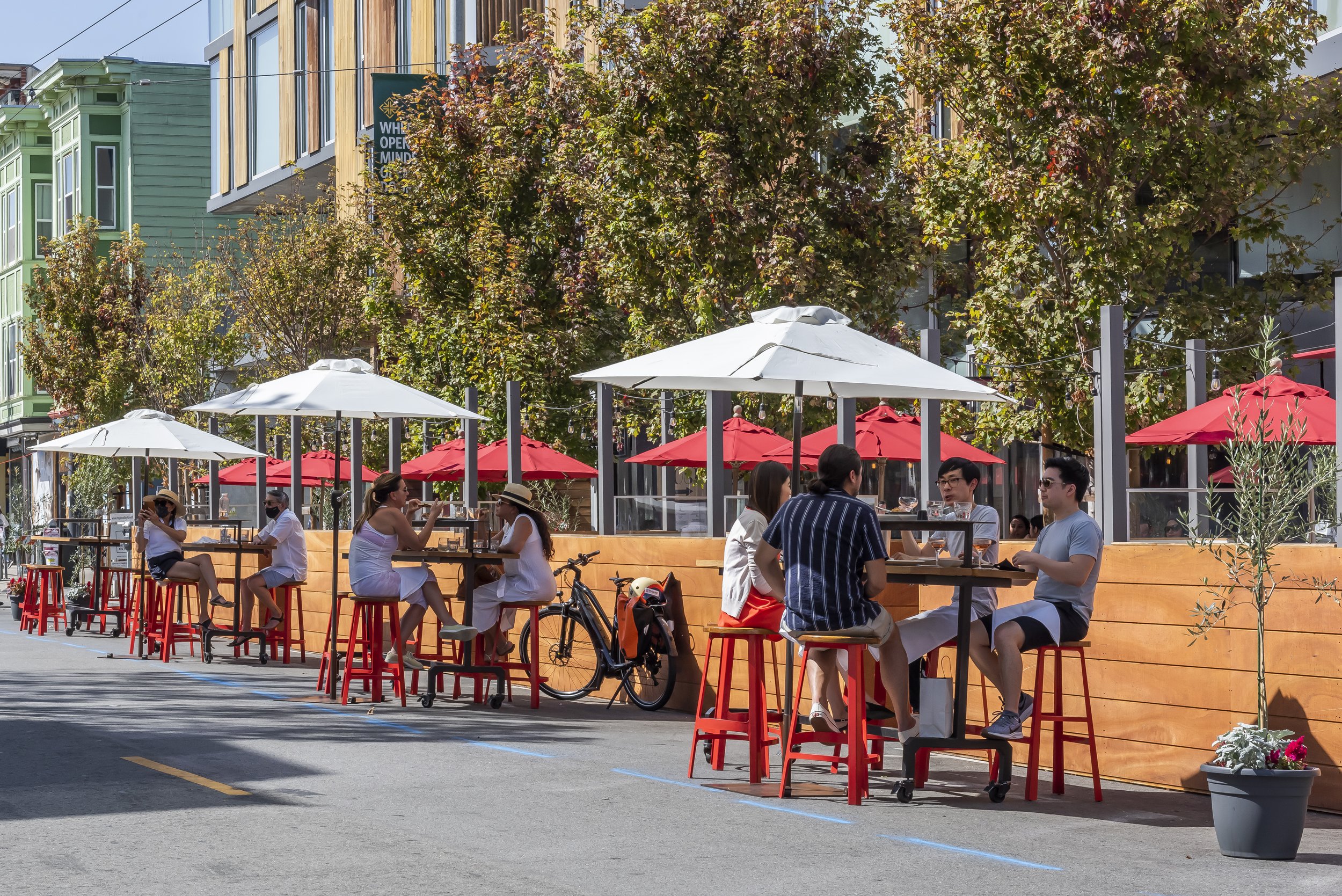 People dining outdoors at a restaurant with red and white umbrellas, in front of colorful buildings and green trees on a sunny day.
