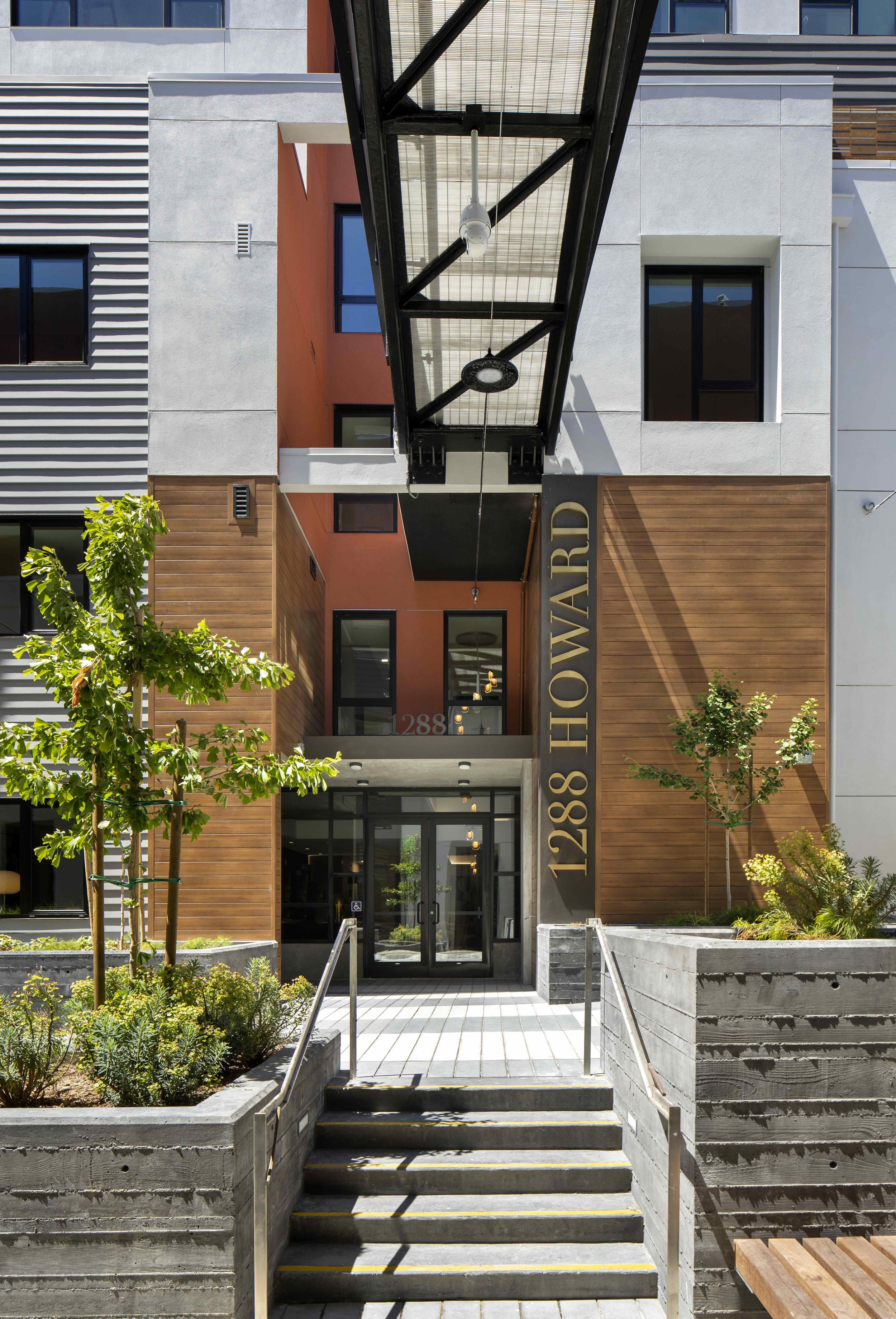 Modern building entrance with steps leading up to glass doors, potted plants, trees, and a large vertical sign with the number 1288 and the word 'HOWARD' on it.