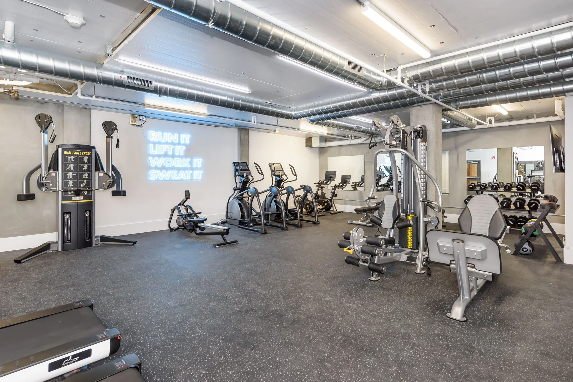 Interior of a modern gym with various workout equipment including treadmills, exercise bikes, weight machines, and dumbbells. There is a neon sign on the wall that says "RUN IT LIFT IT WORK IT SWEAT IT." The ceiling features exposed metal ducts and b