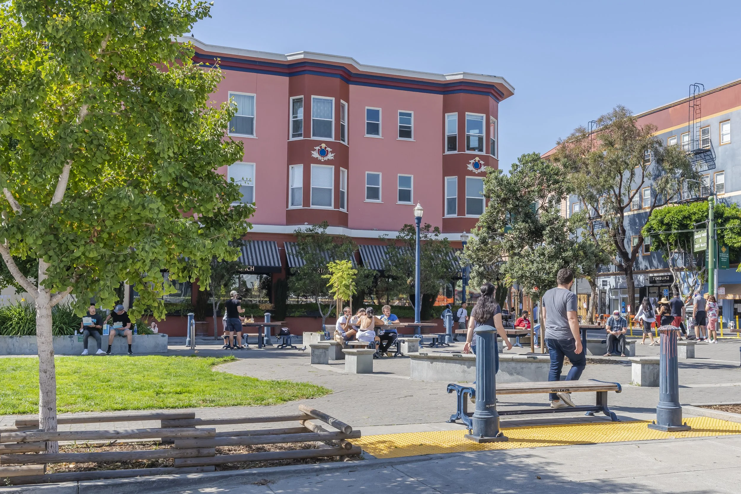 People sitting on benches and walking in a lively park with green trees, pink and blue buildings, and a clear blue sky.