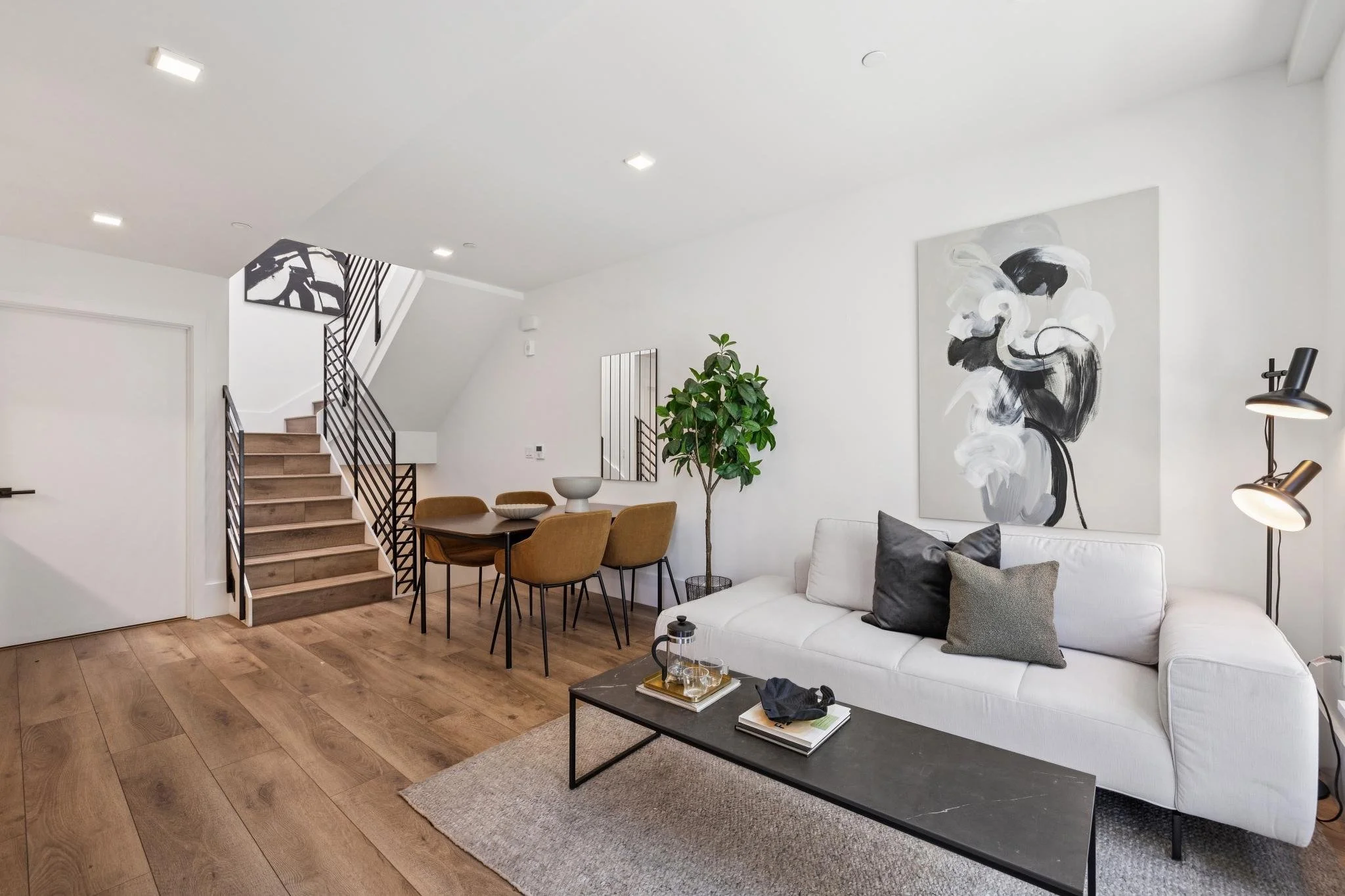 Modern living room with white walls, wooden flooring, a white sofa with gray pillows, a black coffee table with books and glasses, a large abstract black and white painting, a potted plant, a dining table with tan chairs, and stairs with black railin