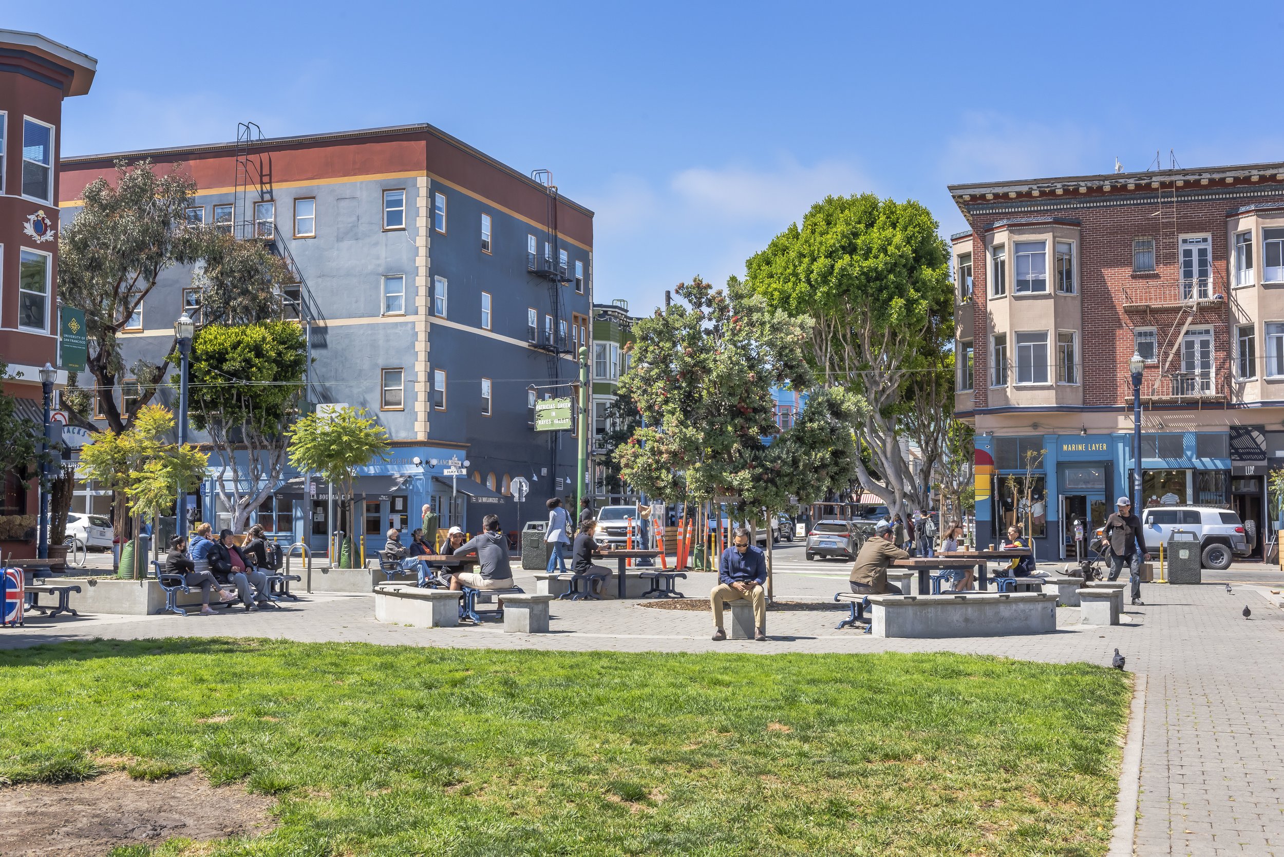 People sitting and walking in a park with benches, trees, and buildings in the background on a sunny day.