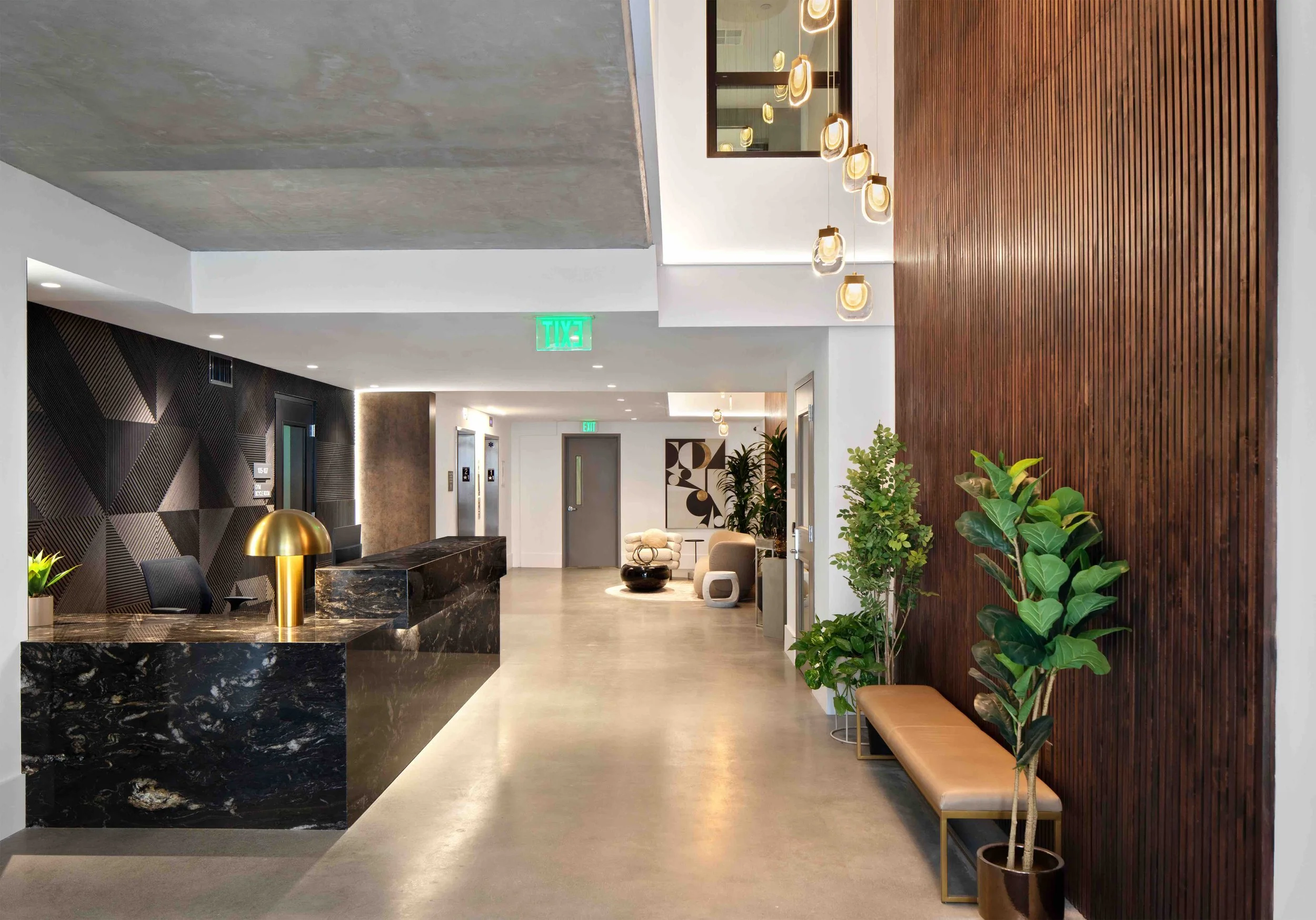 Modern hotel lobby with a black marble reception desk, gold lamp, decorative wall art, and indoor plants, featuring a mix of wood paneling and textured black walls.