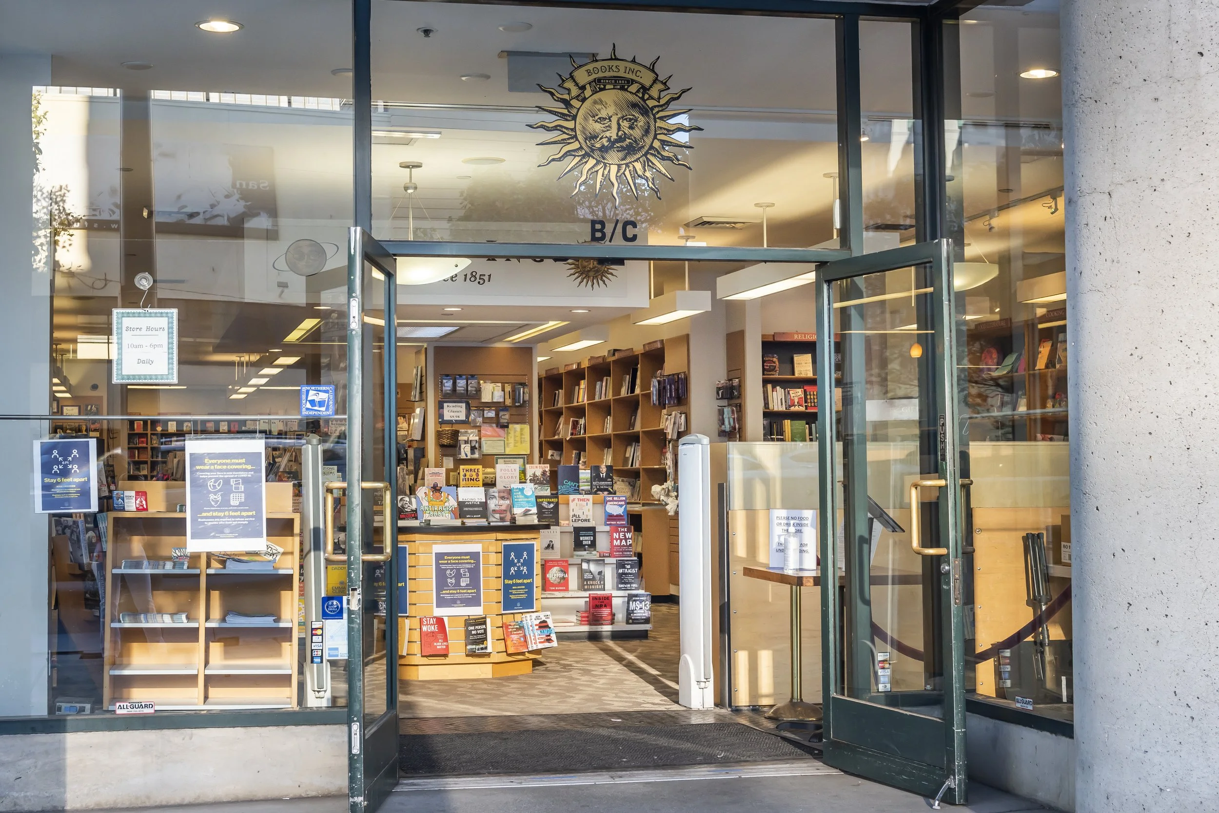 Interior view of a bookstore with shelves filled with books, a display table, and signage, as seen through glass doors, with a logo of a sun with a face and the text "Books Inc." hanging above.