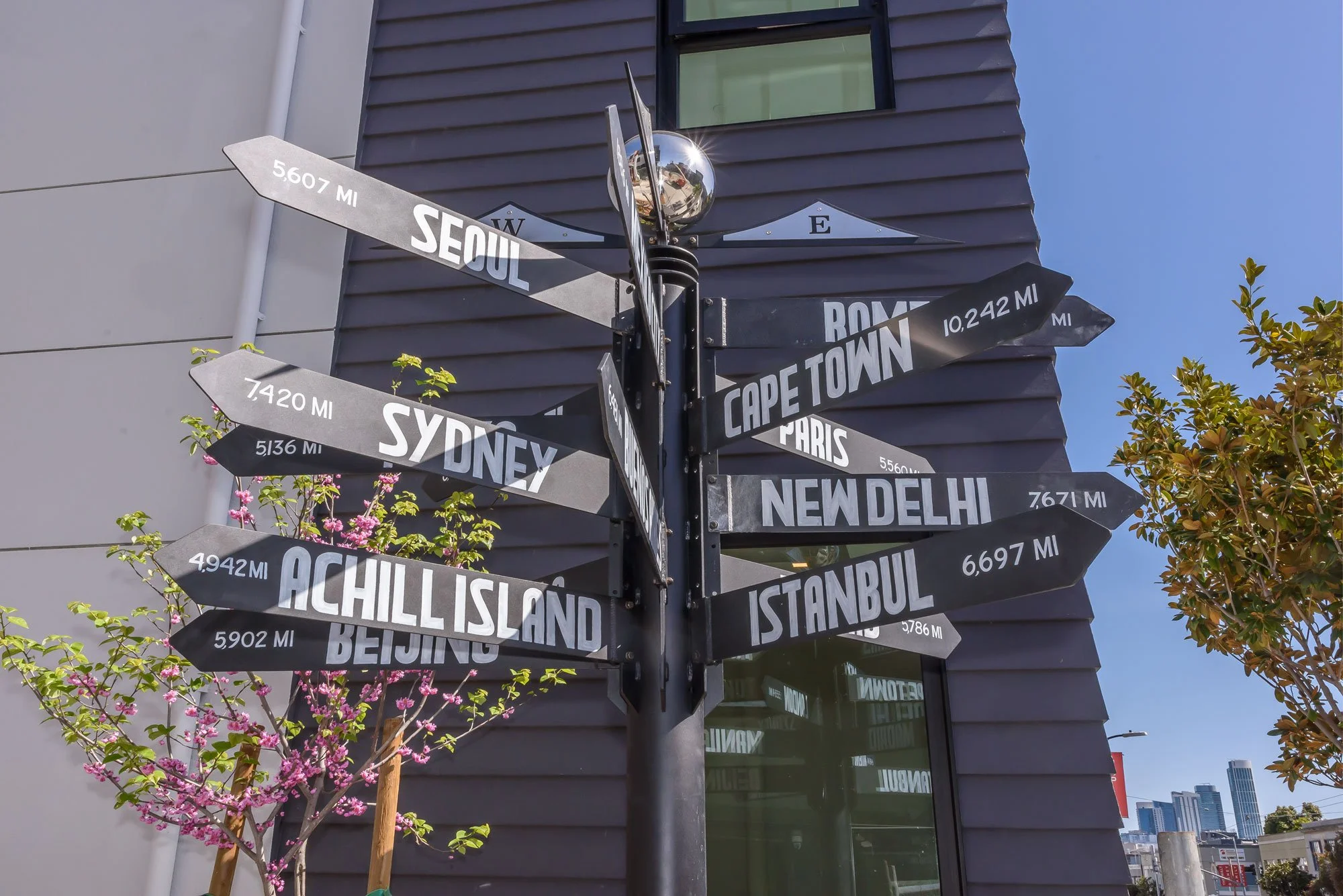 A street signpost with multiple directional signs indicating distances to various global cities, including Seoul, Rome, Cape Town, Sydney, New Delhi, Achill Island, and Istanbul, mounted on a black pole against a modern building with a window, surrou