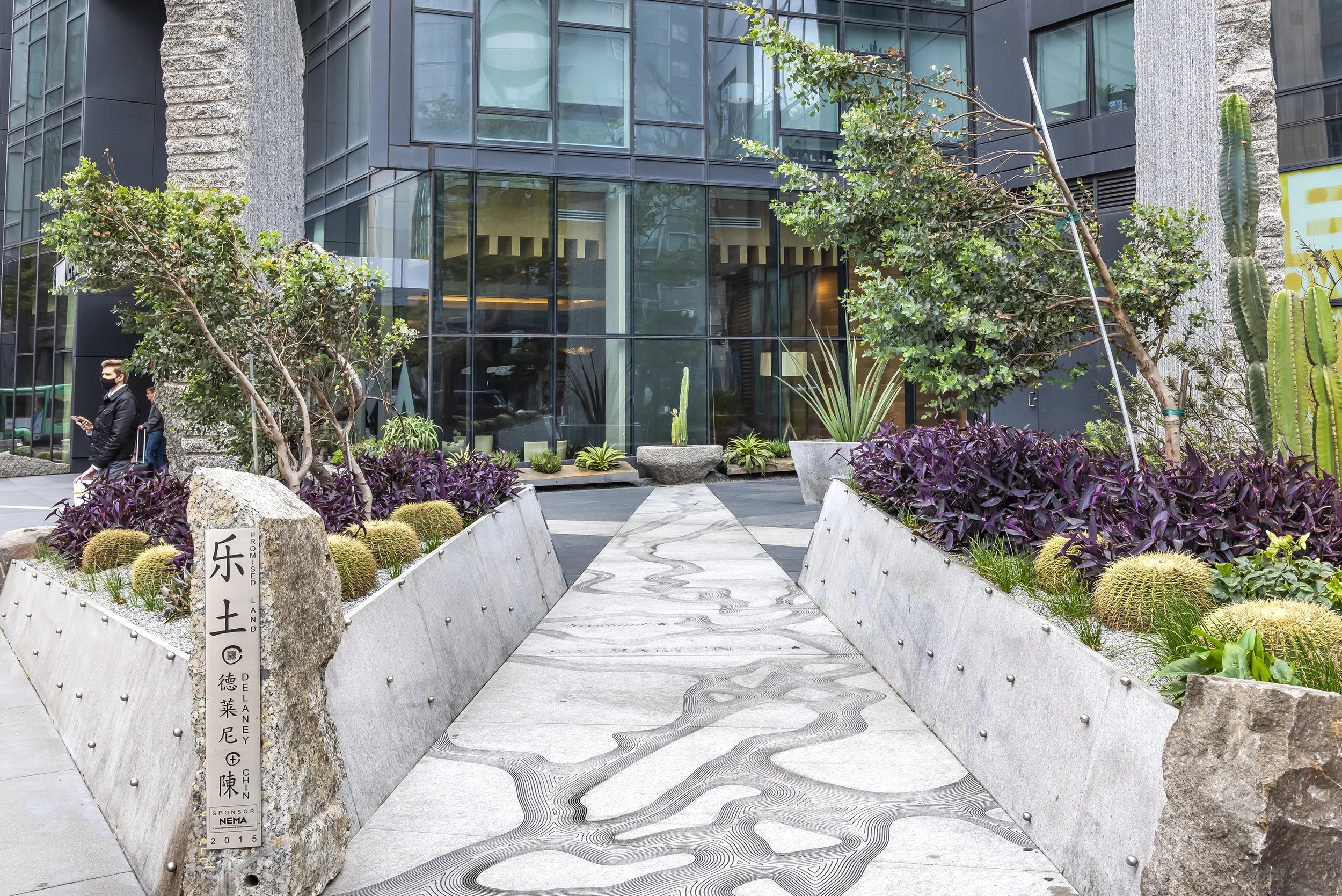 Urban outdoor scene featuring modern glass building facade, landscaped area with purple and green plants, cacti, and people walking, one wearing a face mask, in daytime.