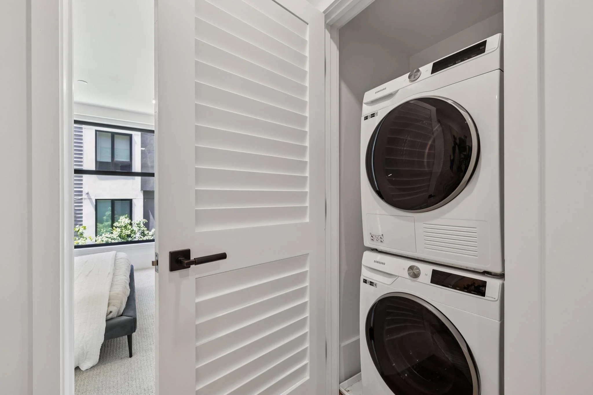A laundry closet with a front-loading washer and dryer stacked on top of each other, a white door with horizontal slats, and a view into a bedroom with a bed and large window.