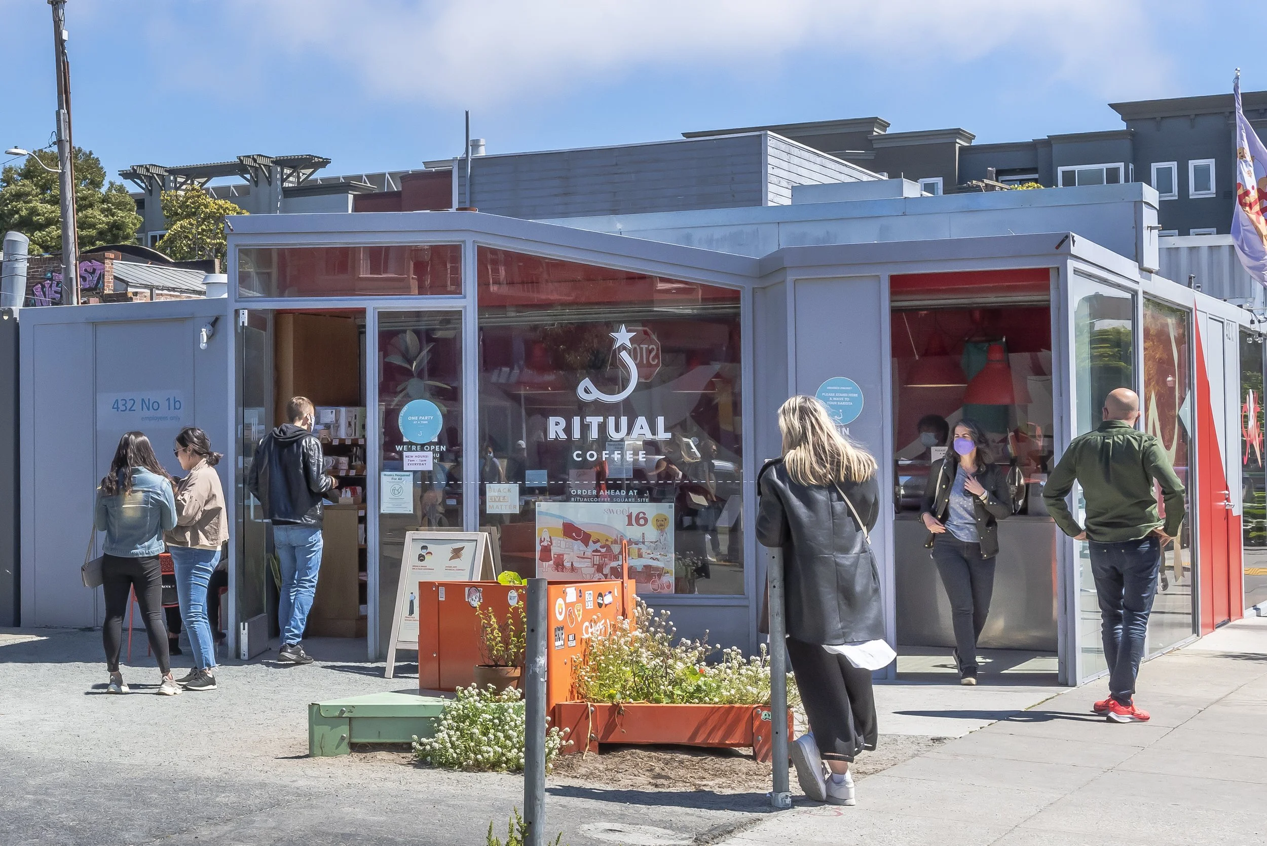 People standing outside a coffee stand called Ritual Coffee in a city setting with buildings, a flag, and flower pots visible.