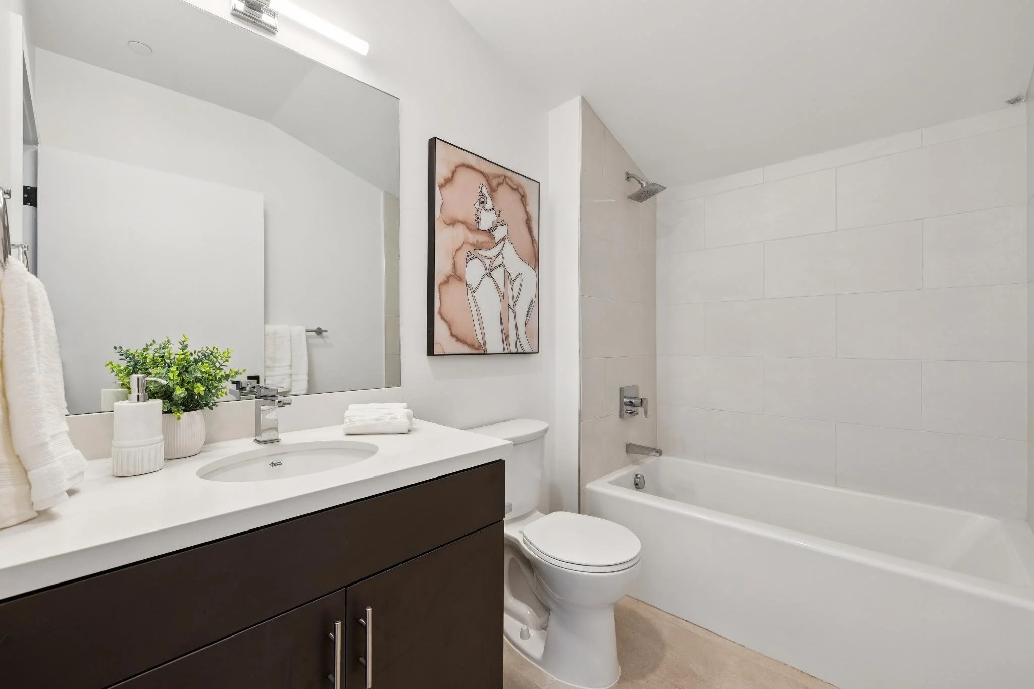 A modern bathroom with a white bathtub, a white toilet, a white sink with a dark cabinet, a large mirror, and wall art of a minimalistic line drawing of a woman. The decor includes towels and a plant on the sink counter.