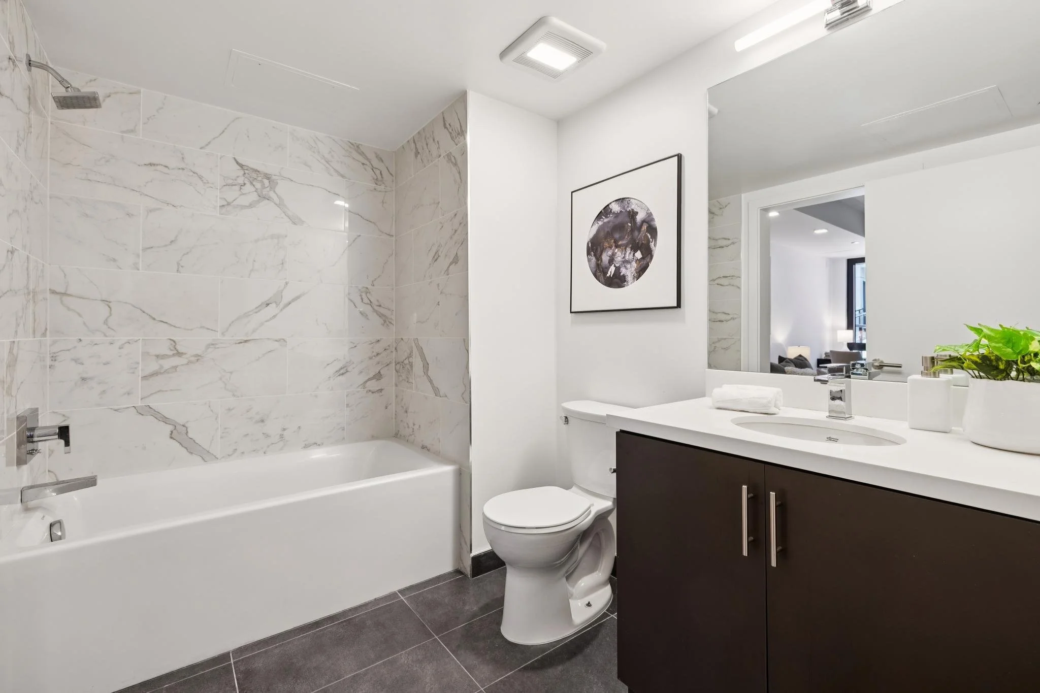 Modern bathroom with a bathtub and marble tiled walls, a white toilet, a dark wooden vanity with a white countertop, a mirror, a framed artwork, a faucet, a potted plant, and a doorway revealing a living room.