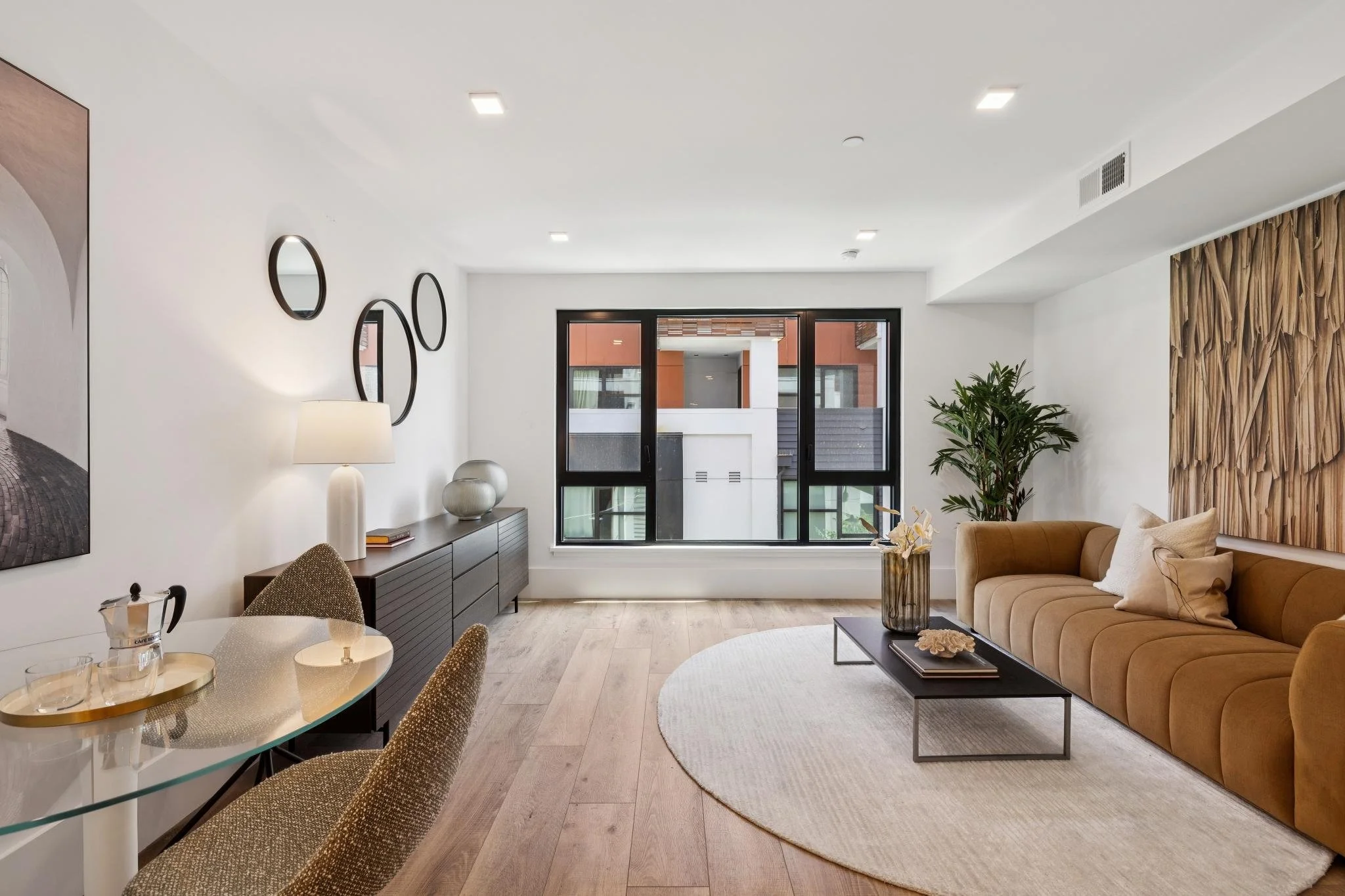 Living room with beige sofa, wooden wall decor, green plant, black coffee table, round white rug, large windows, and modern furniture.