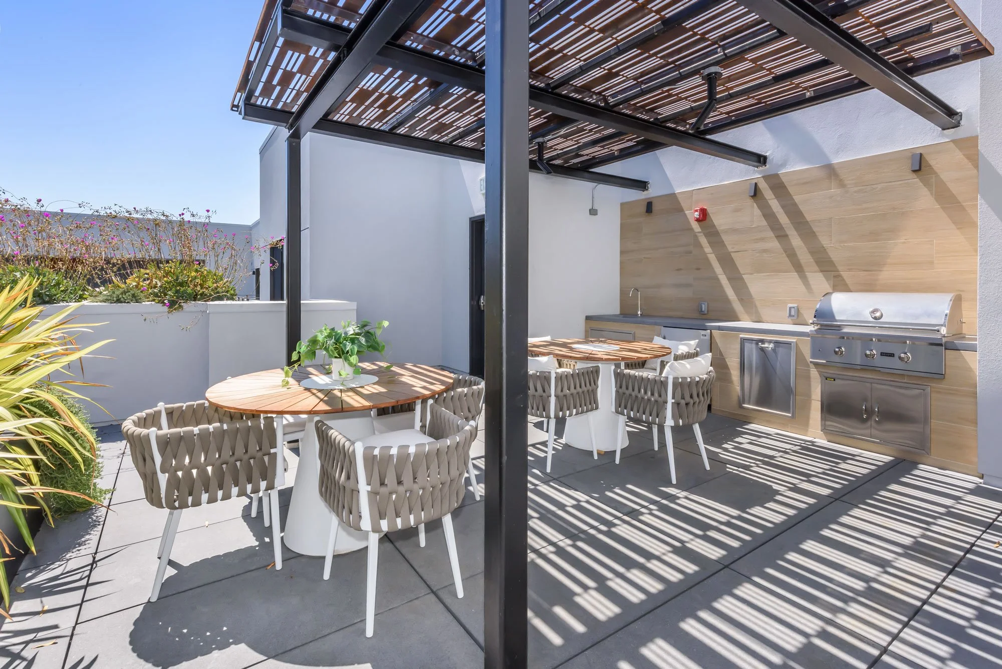 Outdoor patio with a wooden pergola casting striped shadows on the floor, featuring round tables with white chairs, a built-in outdoor kitchen with a grill, sink, and wooden paneling, and plants on the left side with clear blue sky overhead.