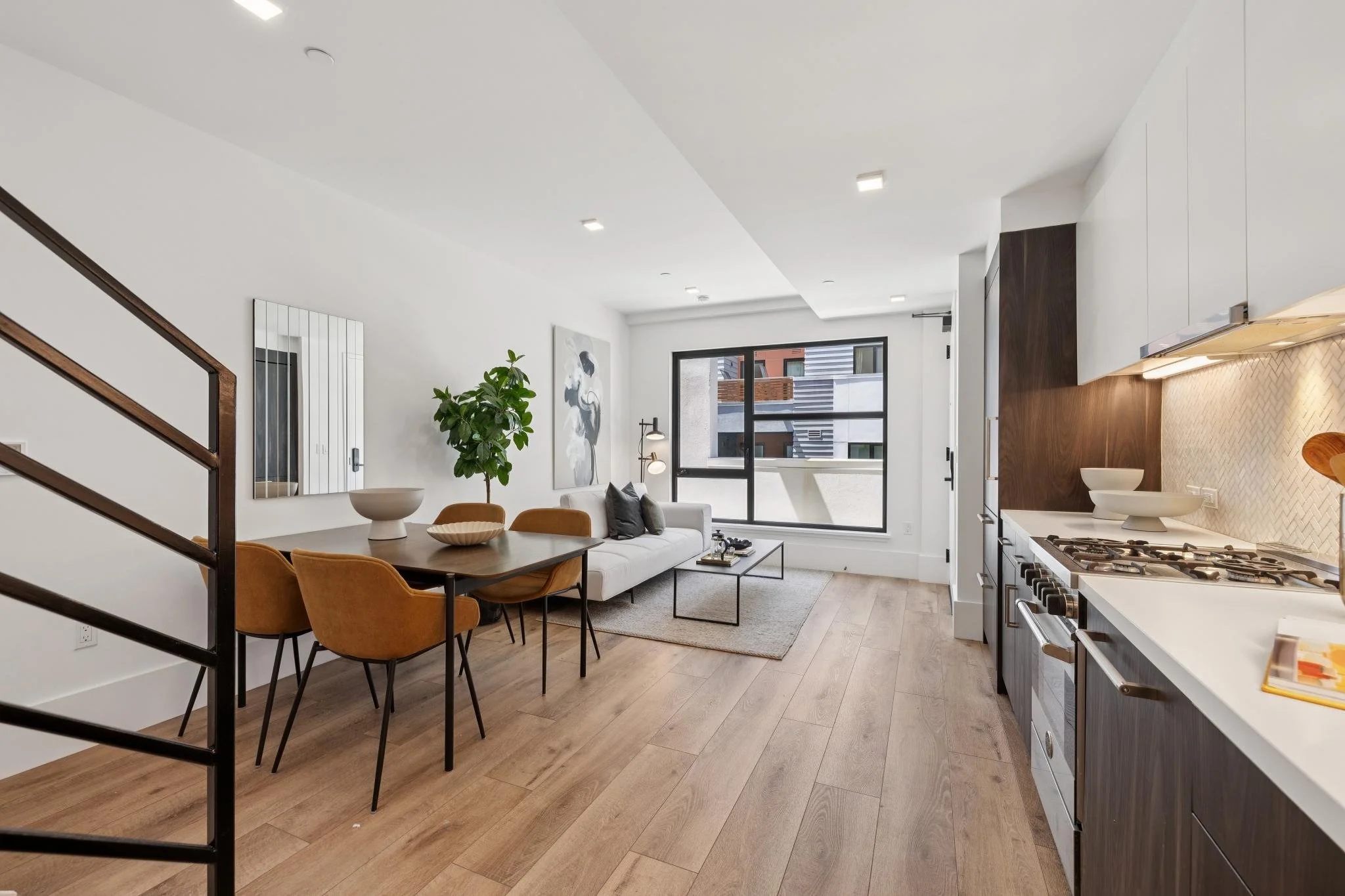 Modern open-plan living room and kitchen with wooden flooring, white walls, a gray sofa, a wooden dining table with orange chairs, a large window, and kitchen cabinets with a gas stove.