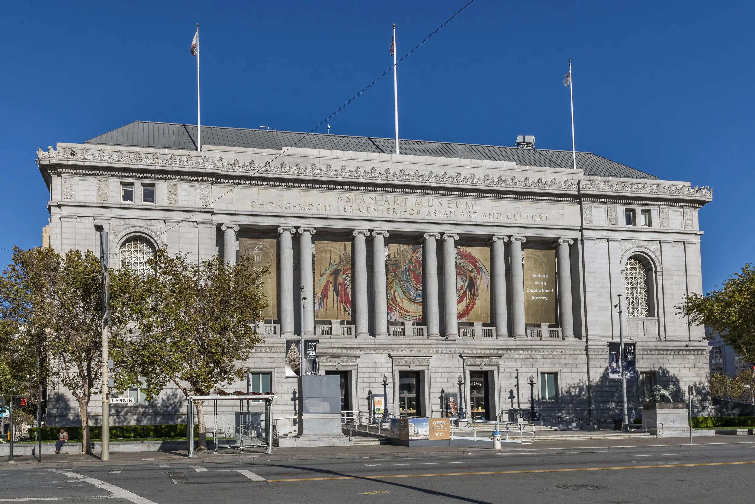 The Chinatown branch of the Asian Art Museum, a neoclassical building with columns, flags on top, and banners on the front, with trees and a street in front.