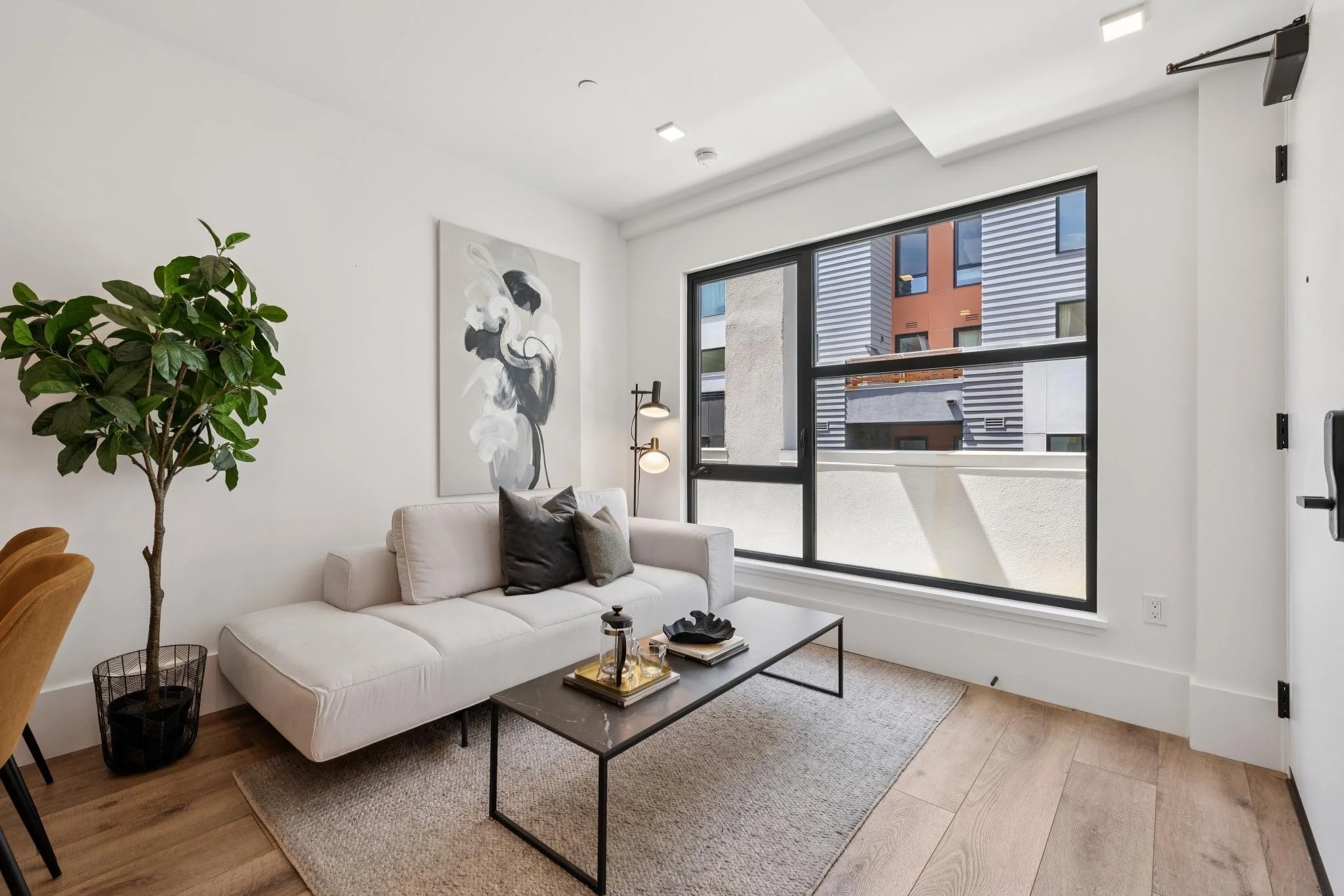 Modern living room with white sofa, black coffee table, large window showing buildings outside, potted plant, abstract wall art, and standing lamp.