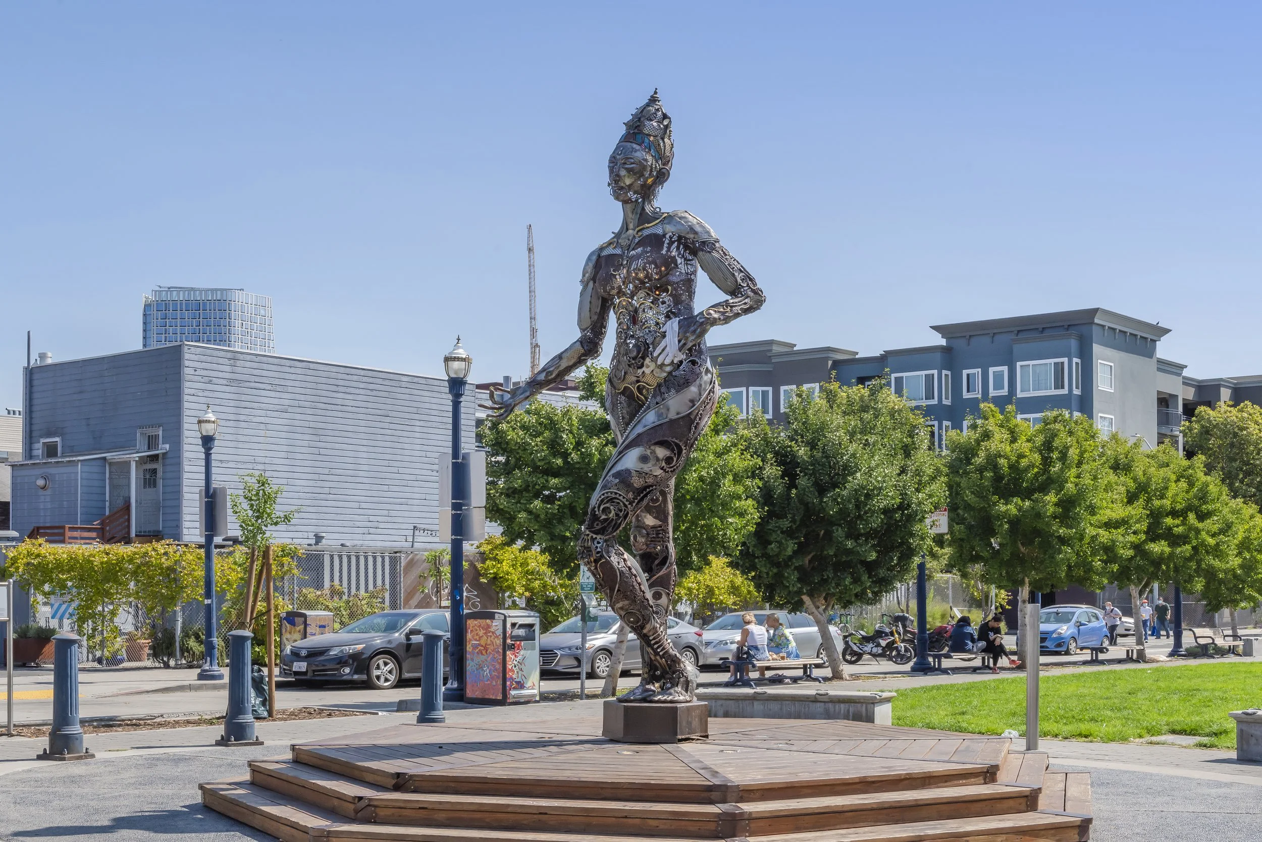 A metal sculpture of a woman standing outdoors in a city park, with buildings and trees in the background.