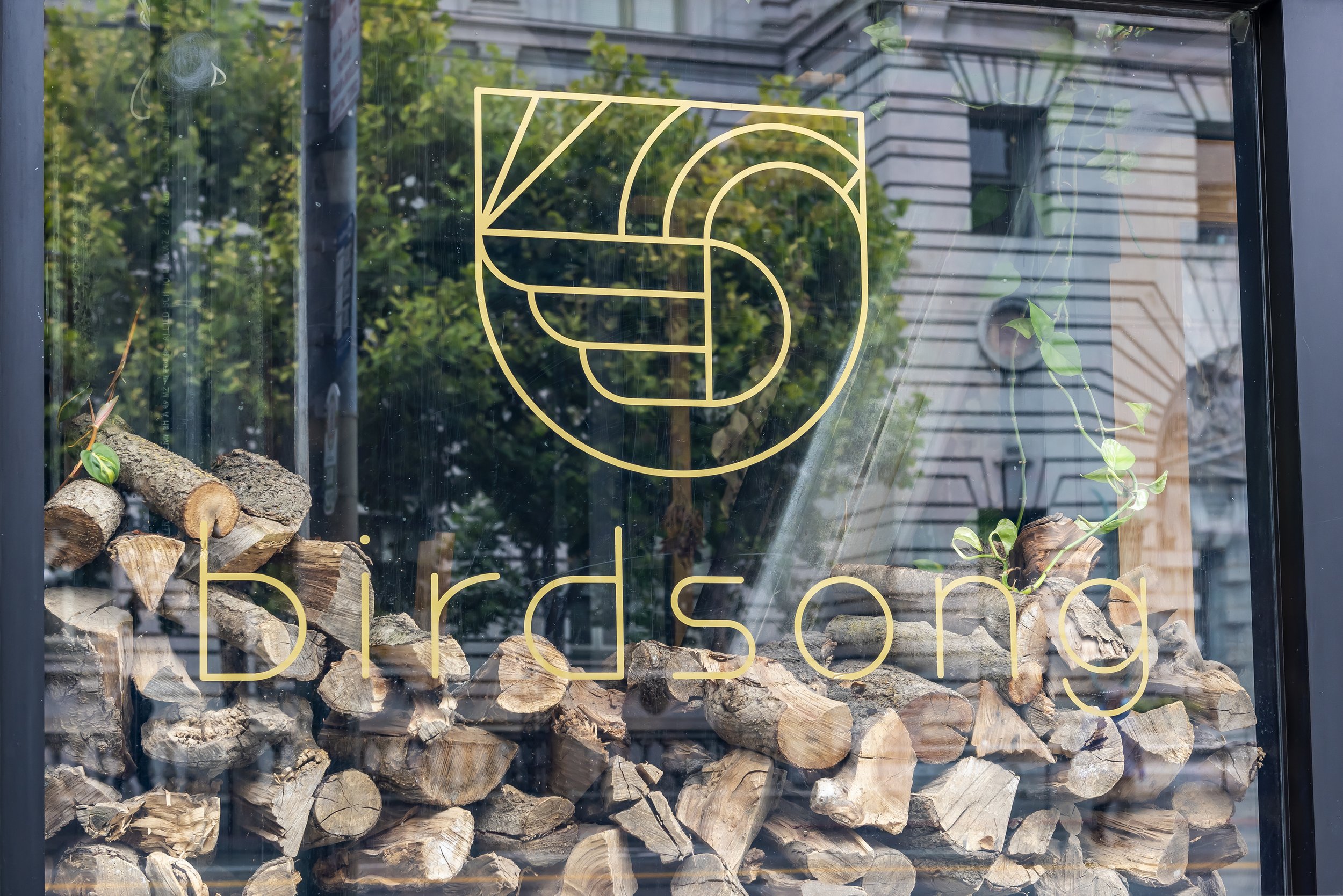 A window display with cut logs piled at the bottom and a gold geometric line drawing of a rose above the word 'garden' on the glass. The background shows trees and a building exterior.
