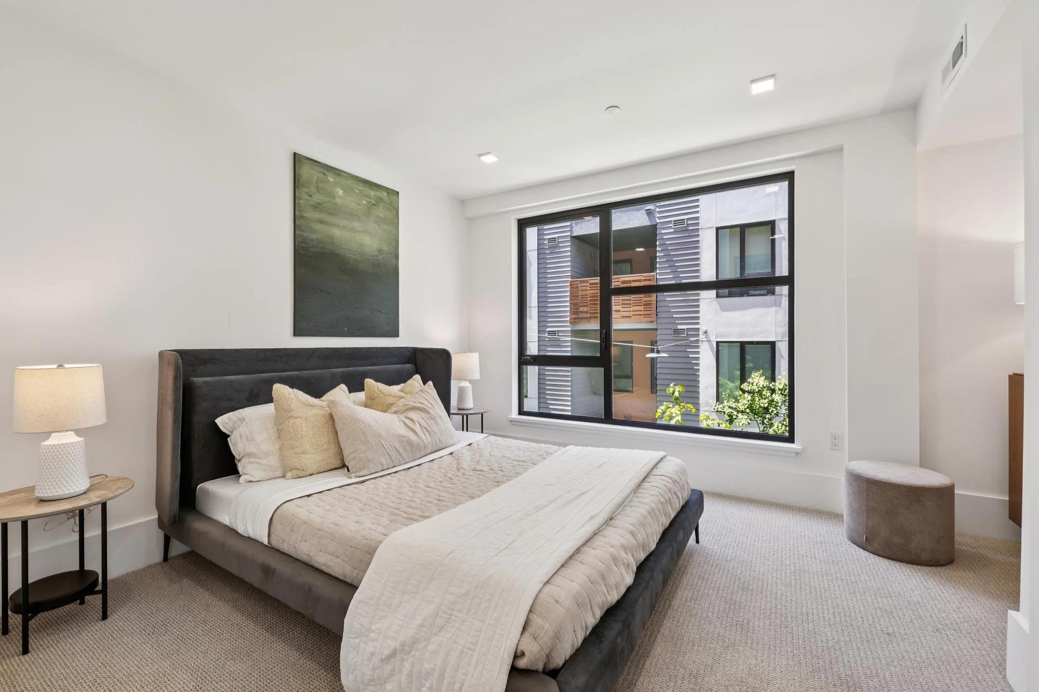 Modern bedroom with a bed, beige bedding, multiple pillows, and bedside lamps, large window with city view, and minimal decor.