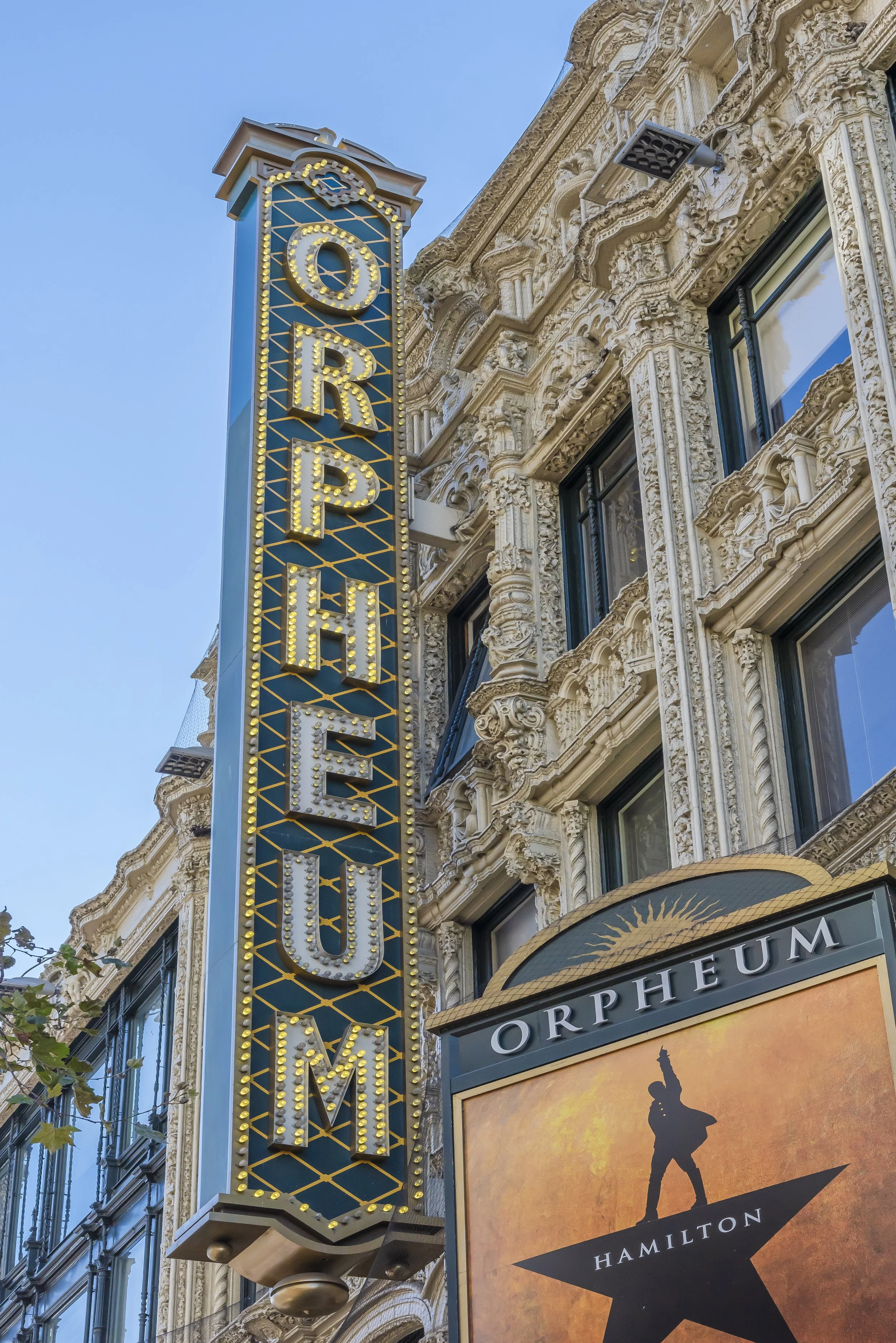 The Orpheum Theatre marquee sign with bright yellow lights and a decorative black and gold background, mounted on an ornate building with elaborate stonework and multiple windows, in an urban setting during daytime.