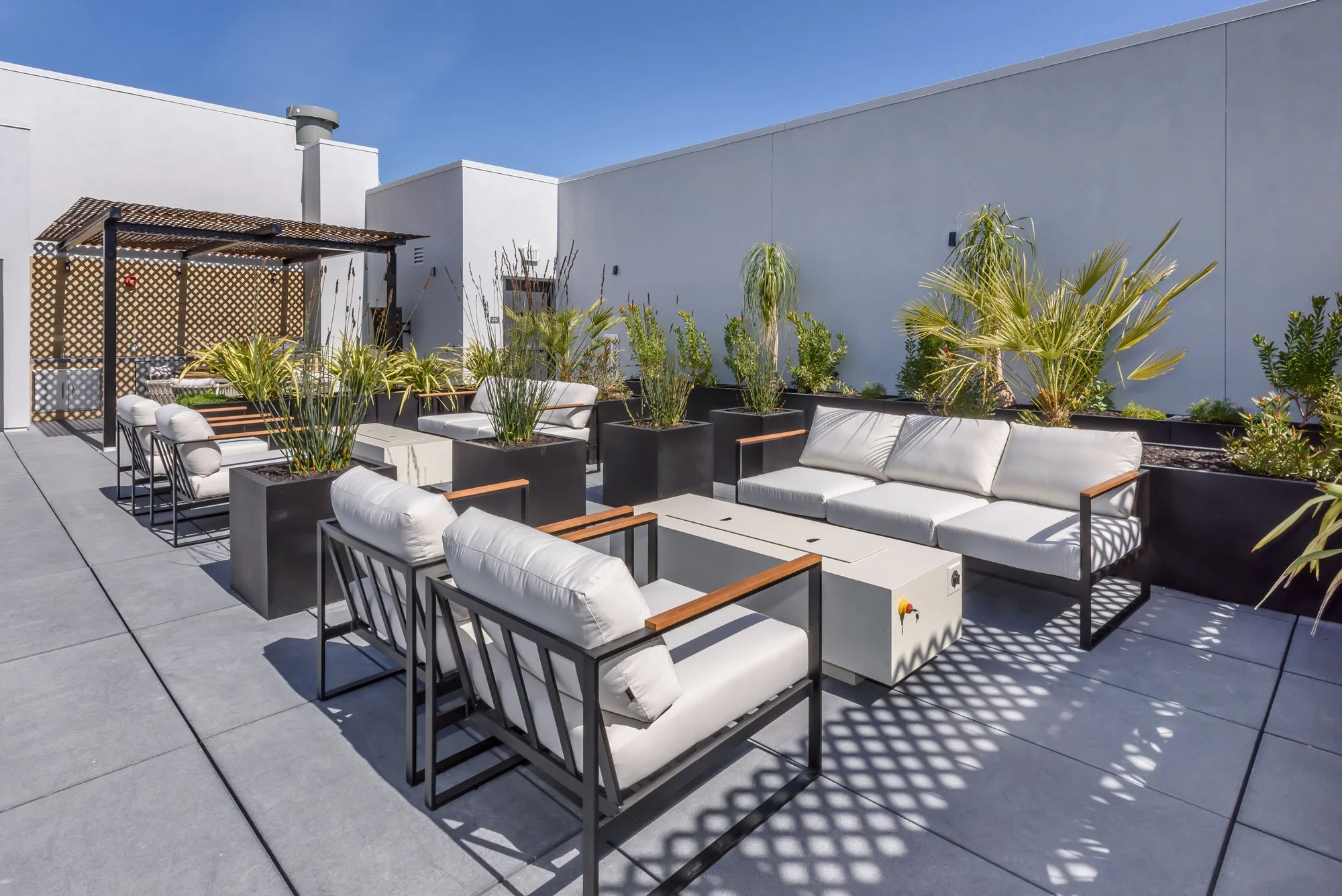 Rooftop patio with white cushioned chairs and sofas, black planters with green plants, wooden lattice structure, and clear blue sky.
