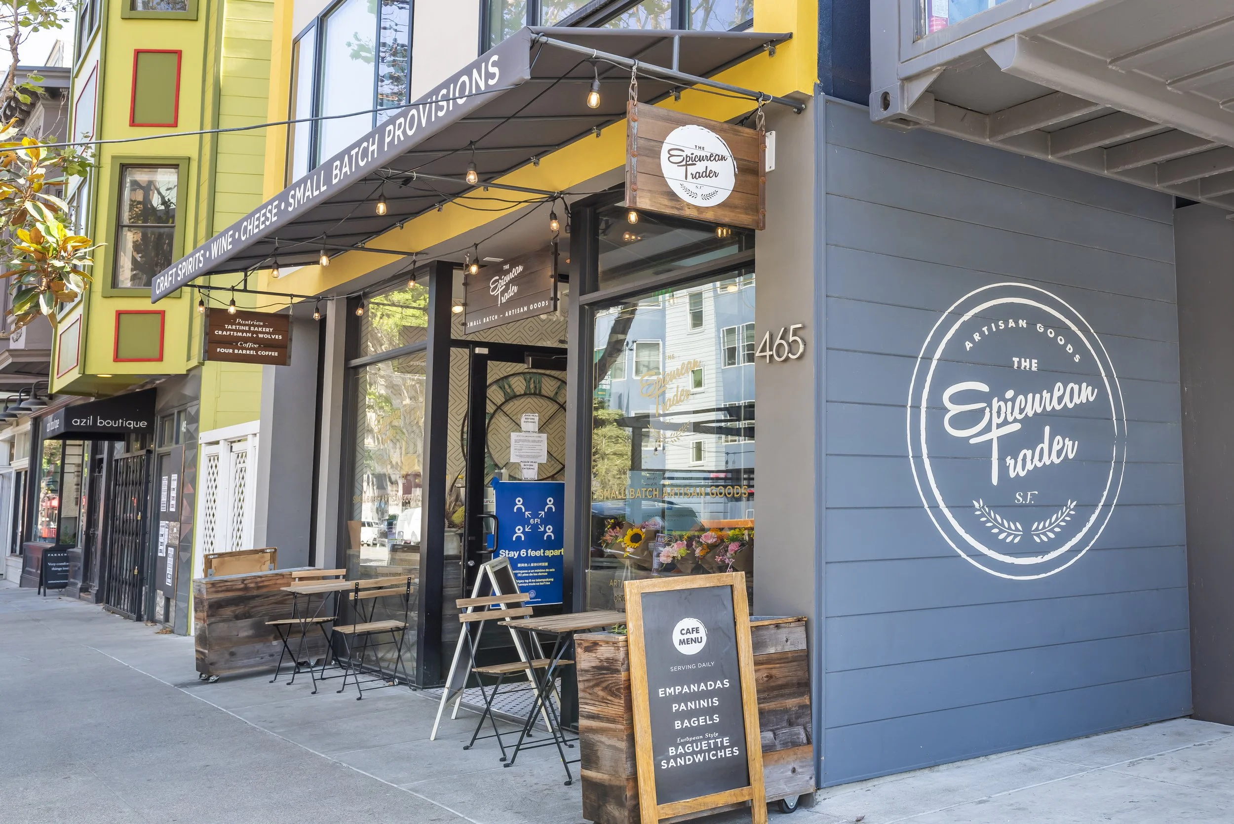 Exterior of The Epicurean Trader, a small artisan goods shop and cafe, with a sidewalk chalkboard menu and outdoor seating on the sidewalk.