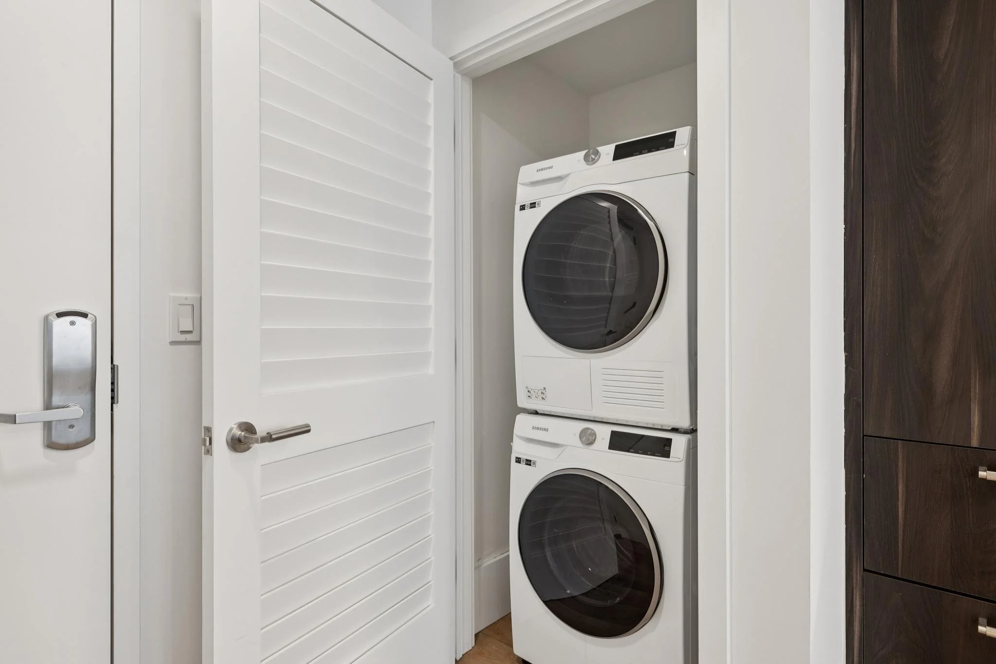 A laundry room with a white door, louvered closet door, and stacked front-loading washer and dryer.