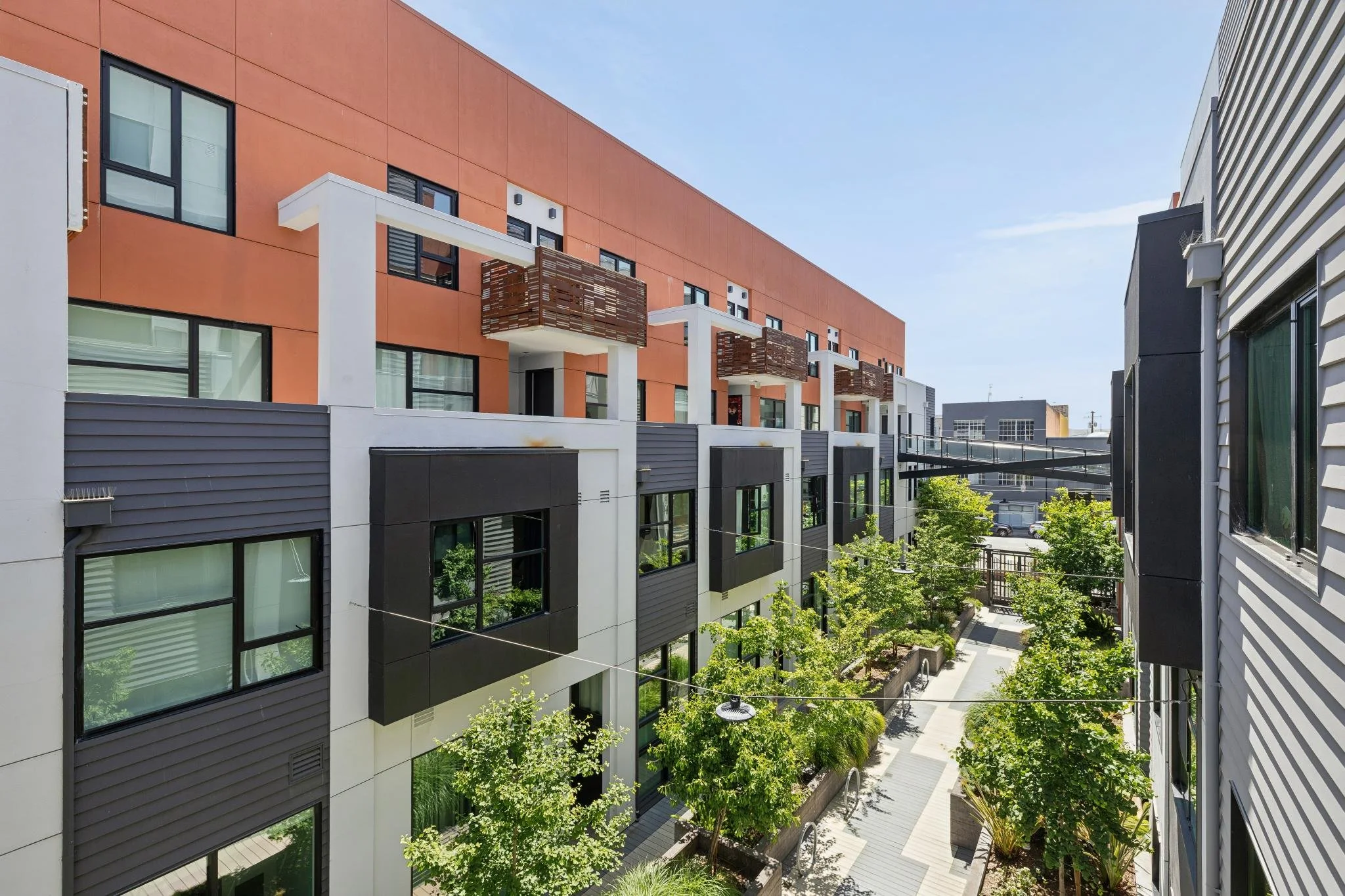 View of modern residential apartment buildings with large windows, balconies with wooden panels, trees, and a clear blue sky.
