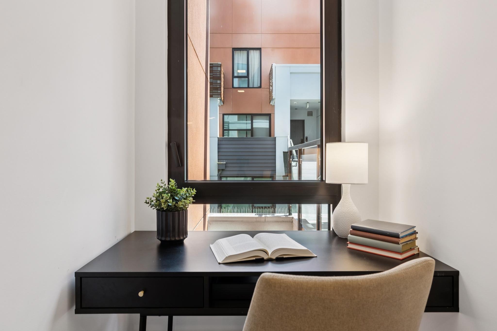 A modern home office with a black desk, beige upholstered chair, and a large window showing an apartment building exterior with pinkish-brown walls and black-framed windows.
