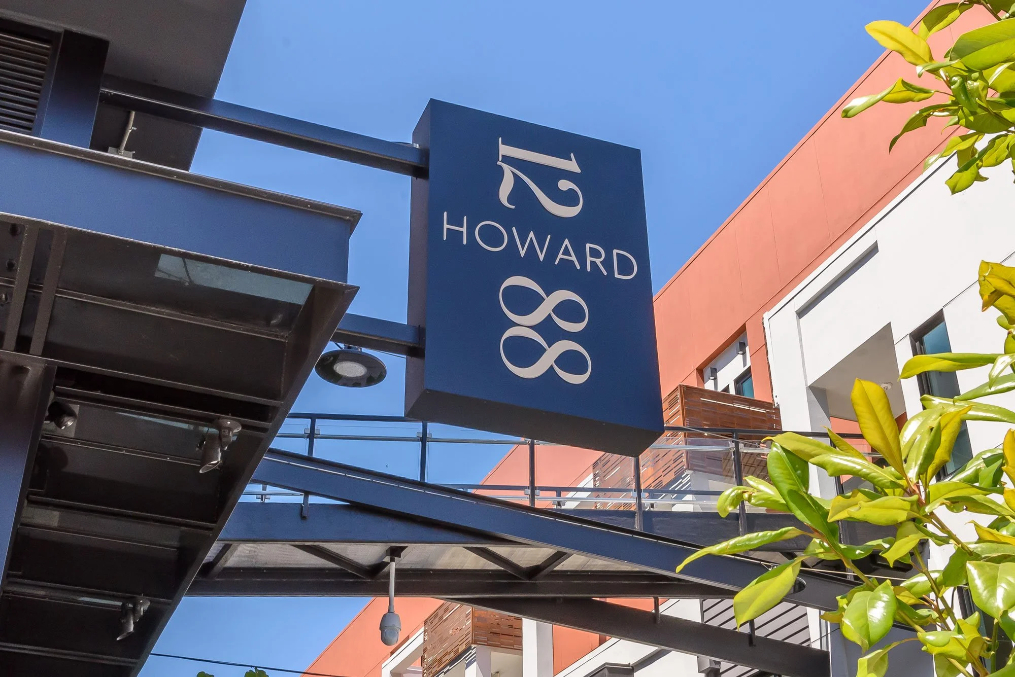 Outdoor shopping mall with a blue sign displaying 'Howard 88' against a clear blue sky, surrounded by modern buildings and greenery.